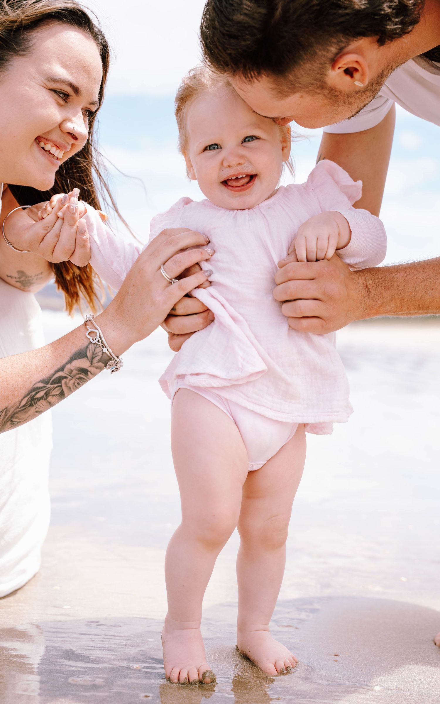 A joyful moment at the beach as parents help their baby in pink take first steps in the sand.