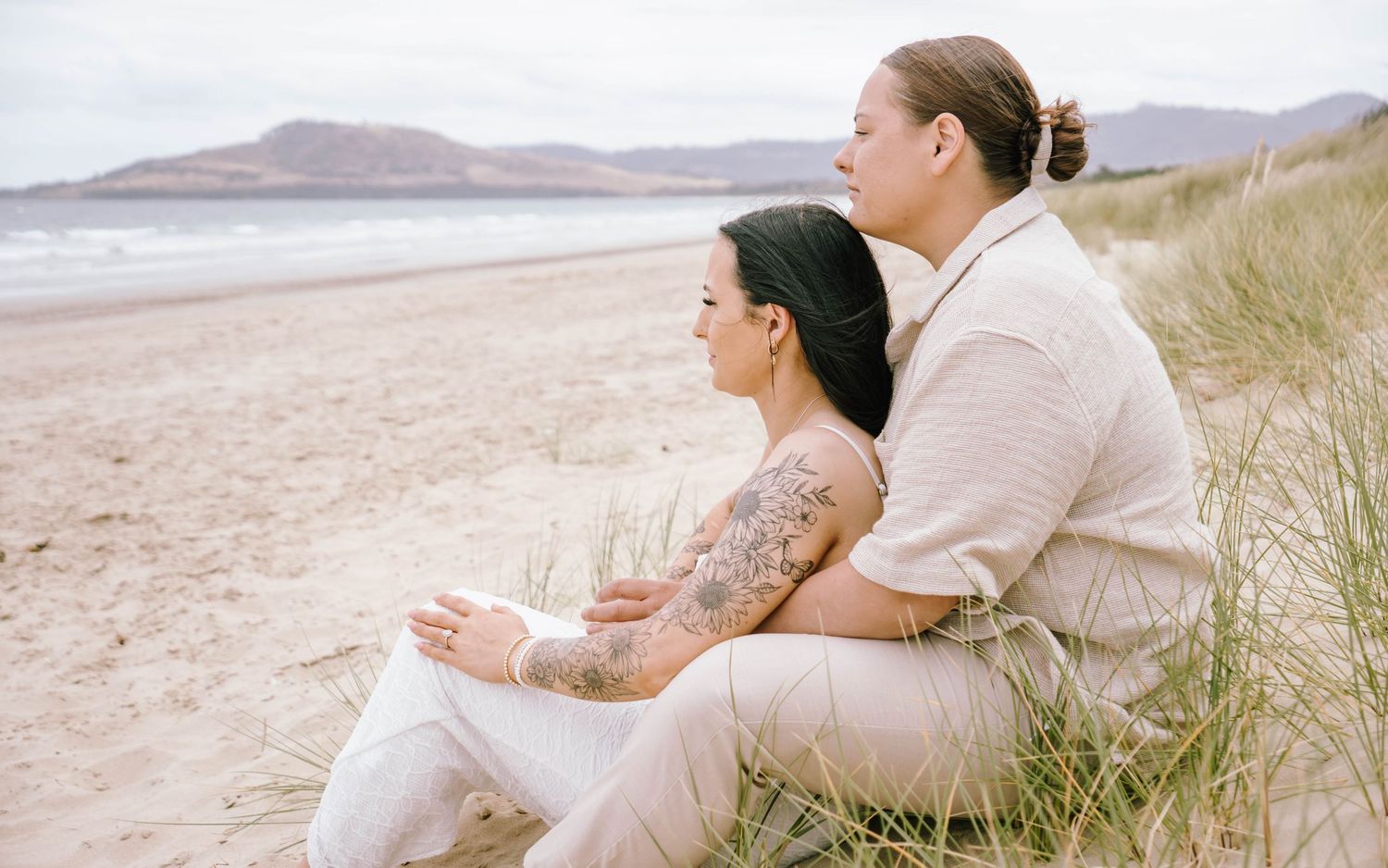 A couple sits together on a sandy beach holding wine glasses while enjoying a peaceful coastal view.