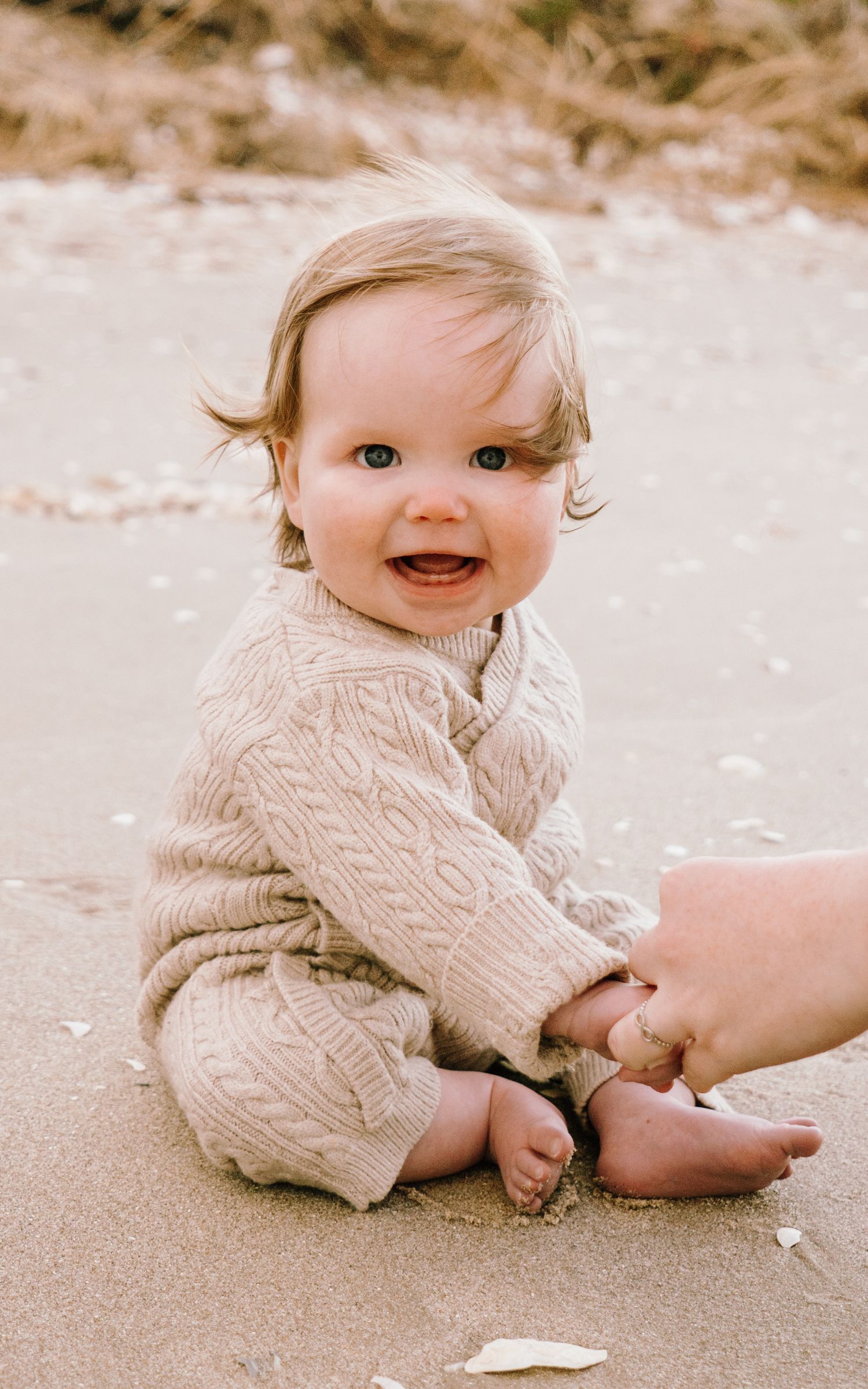 A baby in a cream knit sweater sits on sand and plays with petals in a series of candid photos.