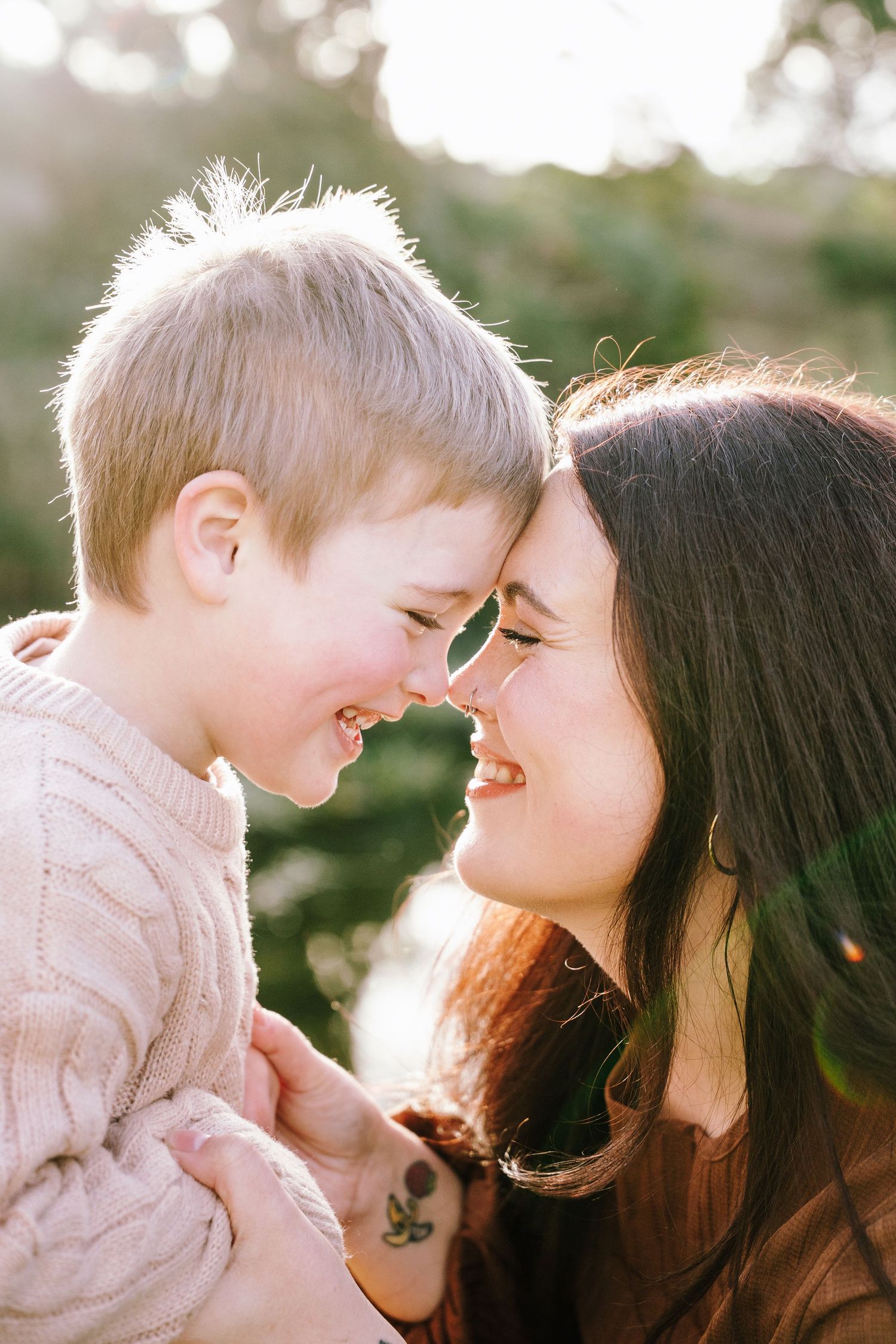 A smiling mother and young child touching noses outdoors in warm golden sunlight, sharing a joyful moment together.