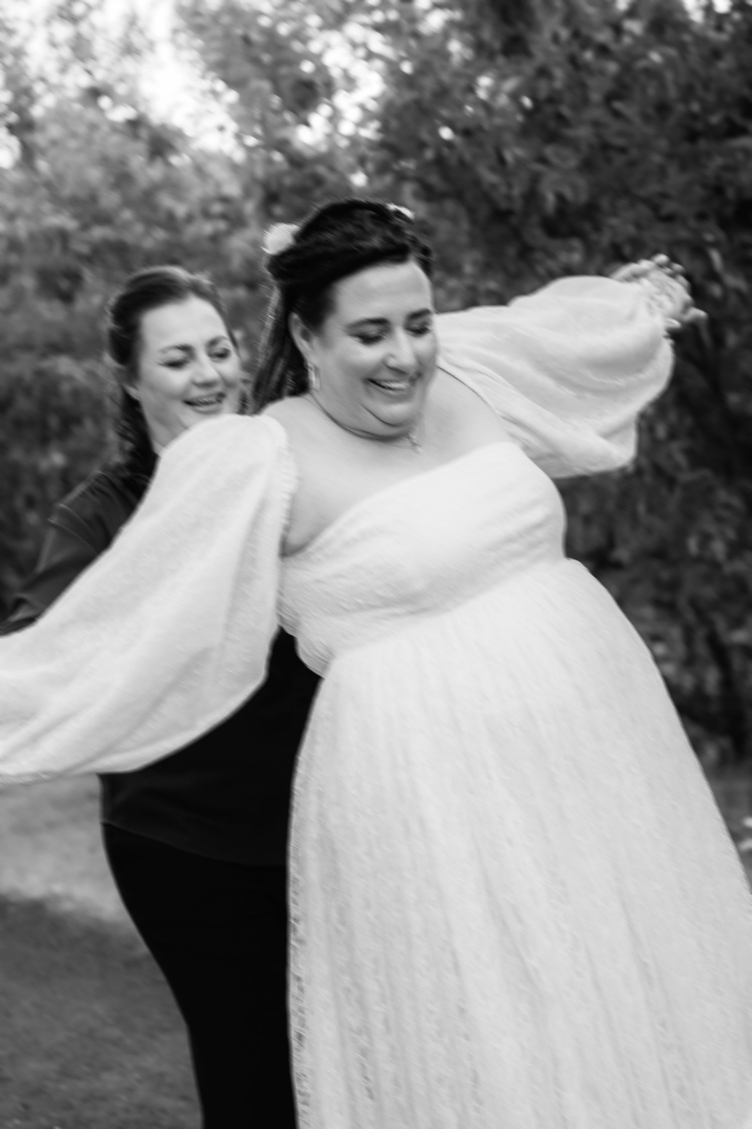 Black and white photo of a bride in a white gown with puff sleeves, joyfully twirling with a woman in black.