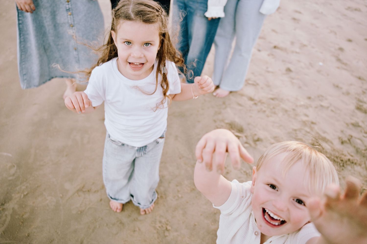 Two young children laughing and reaching toward camera on sandy beach during family photo session.
