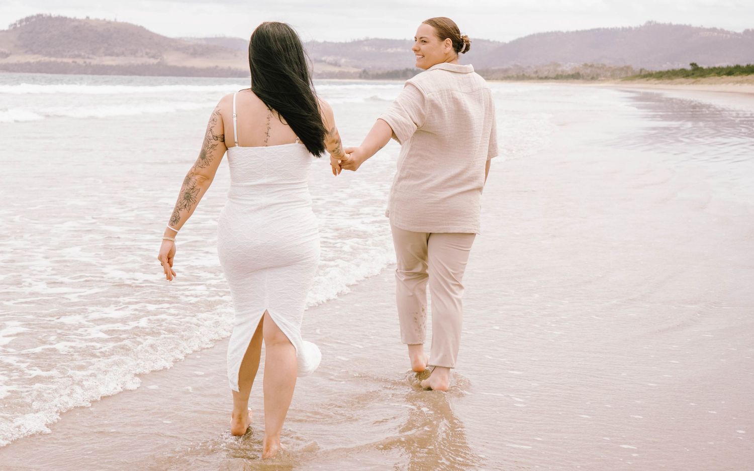 Couple walking hand in hand along a misty beach shoreline in light colored outfits.