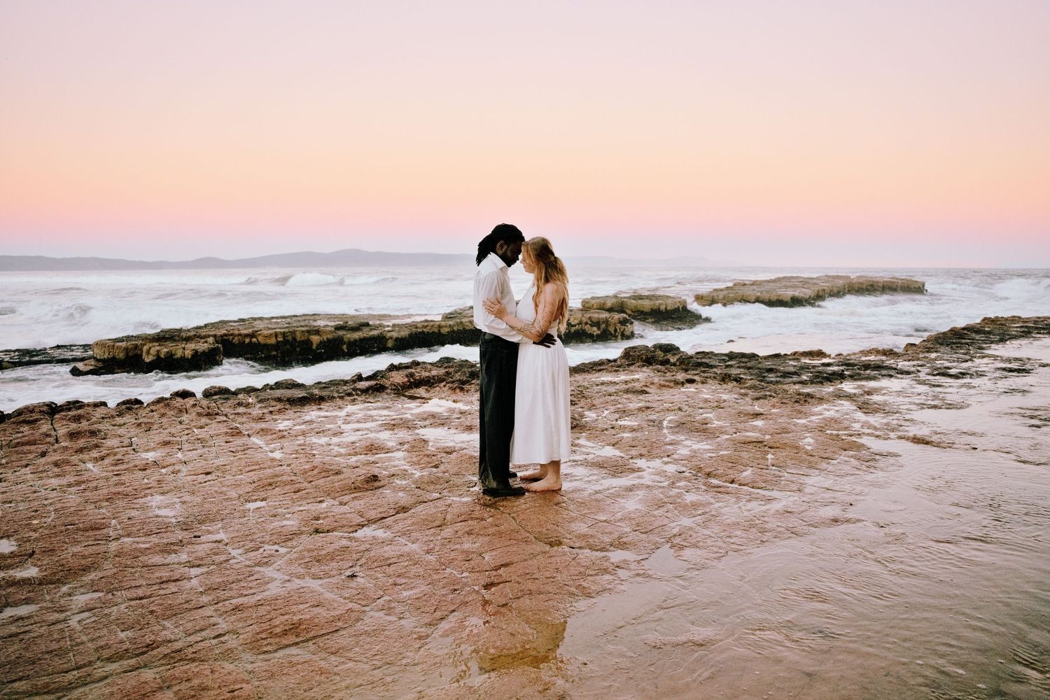 Couple embracing on rocky beach at sunset with pink sky and ocean waves in background.