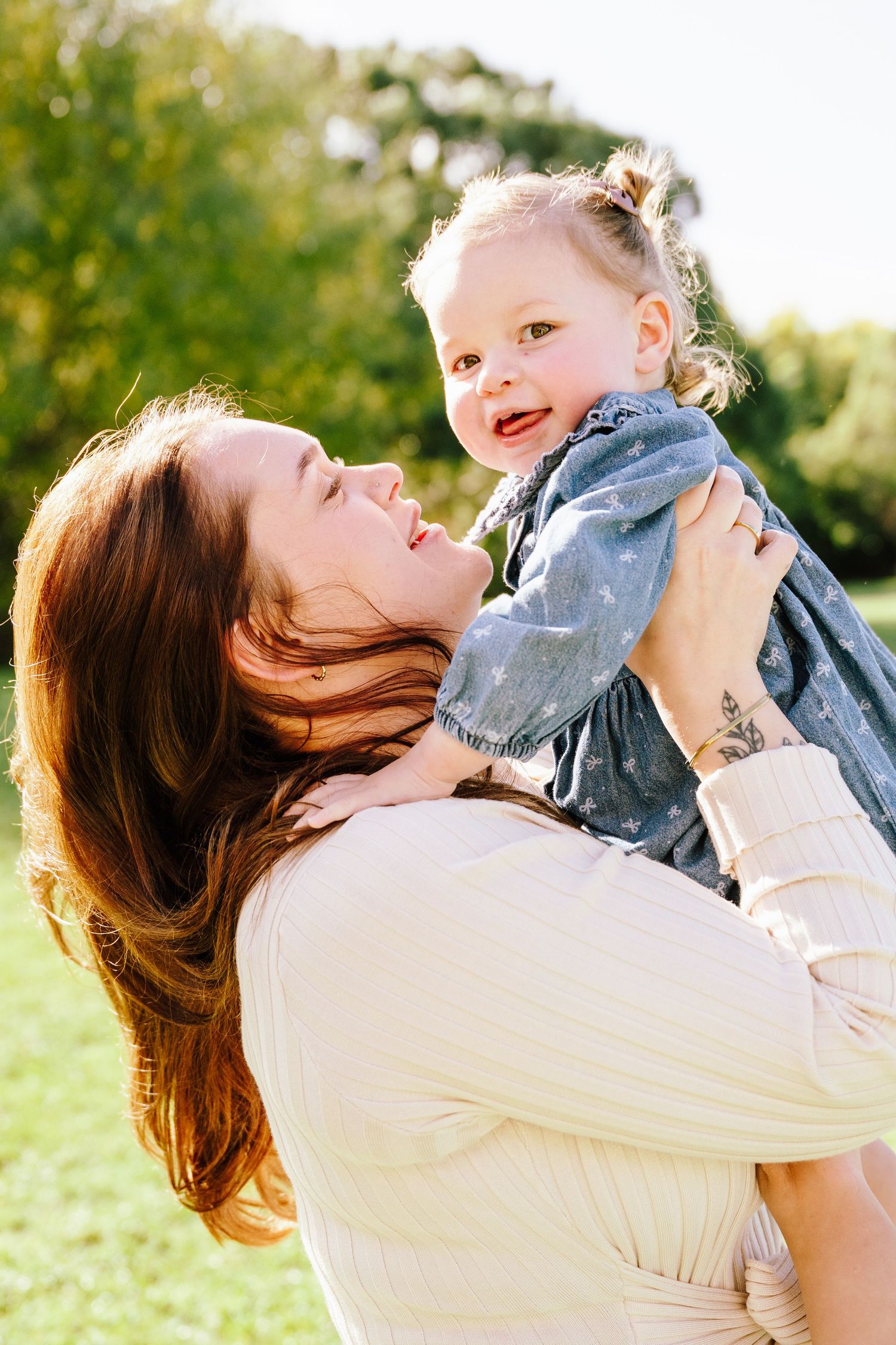 Mother lifts smiling toddler girl in blue dress outdoors in sunny park.