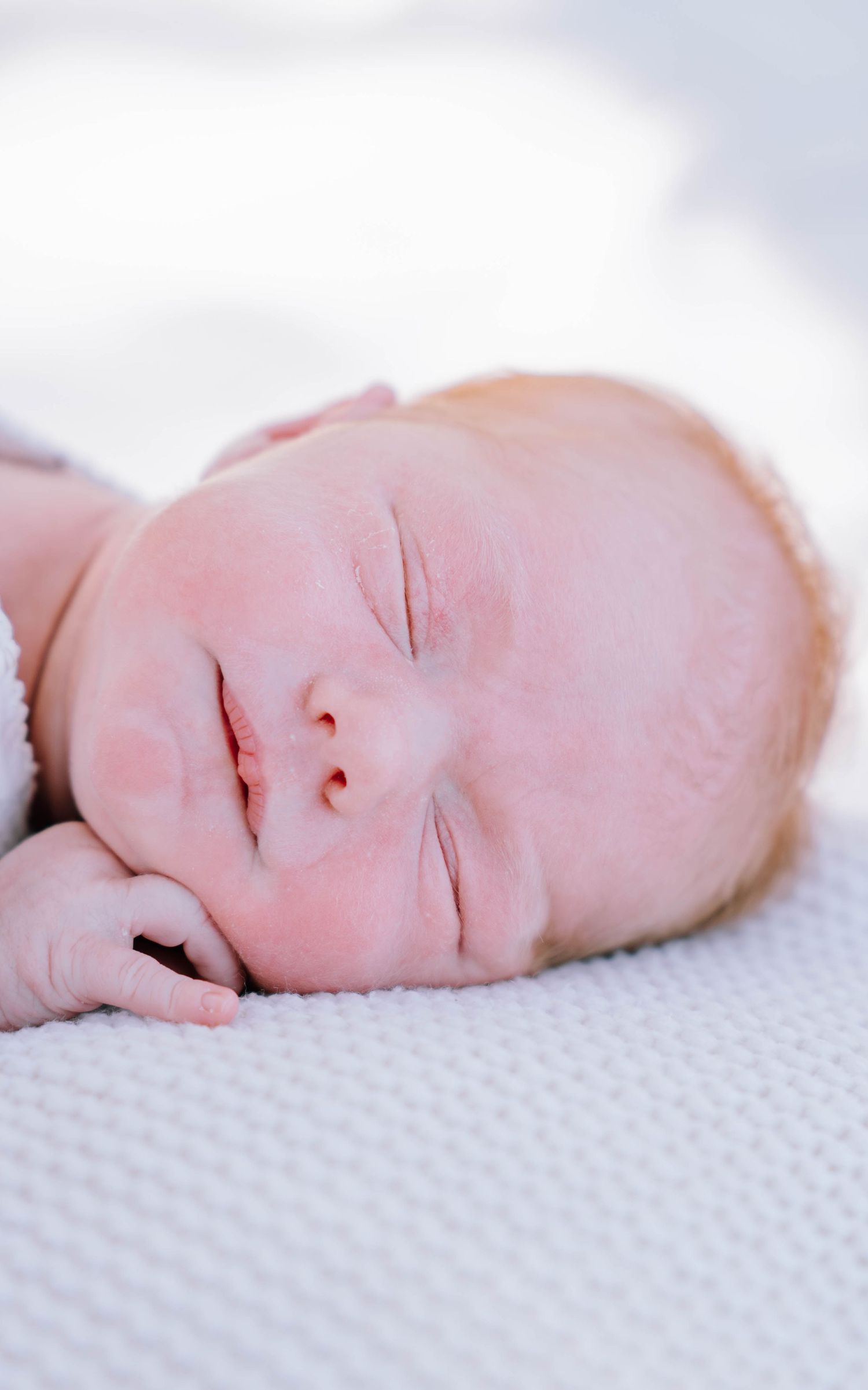 Peaceful newborn baby sleeping on a white knit blanket, with tiny hands near face in soft natural light.