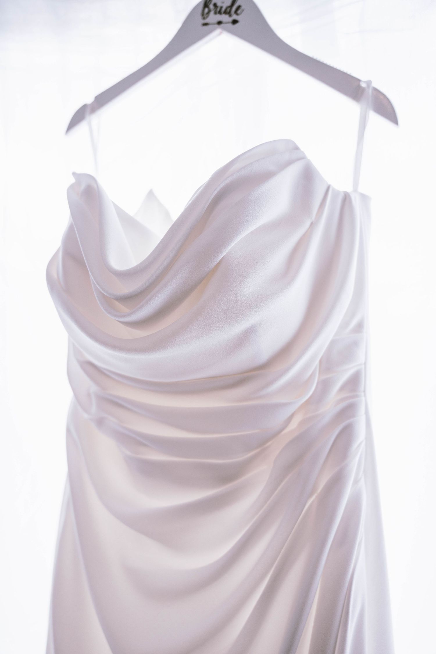 Elegant ivory satin wedding dress with draped cowl neckline hanging on a bridal hanger against a white background.