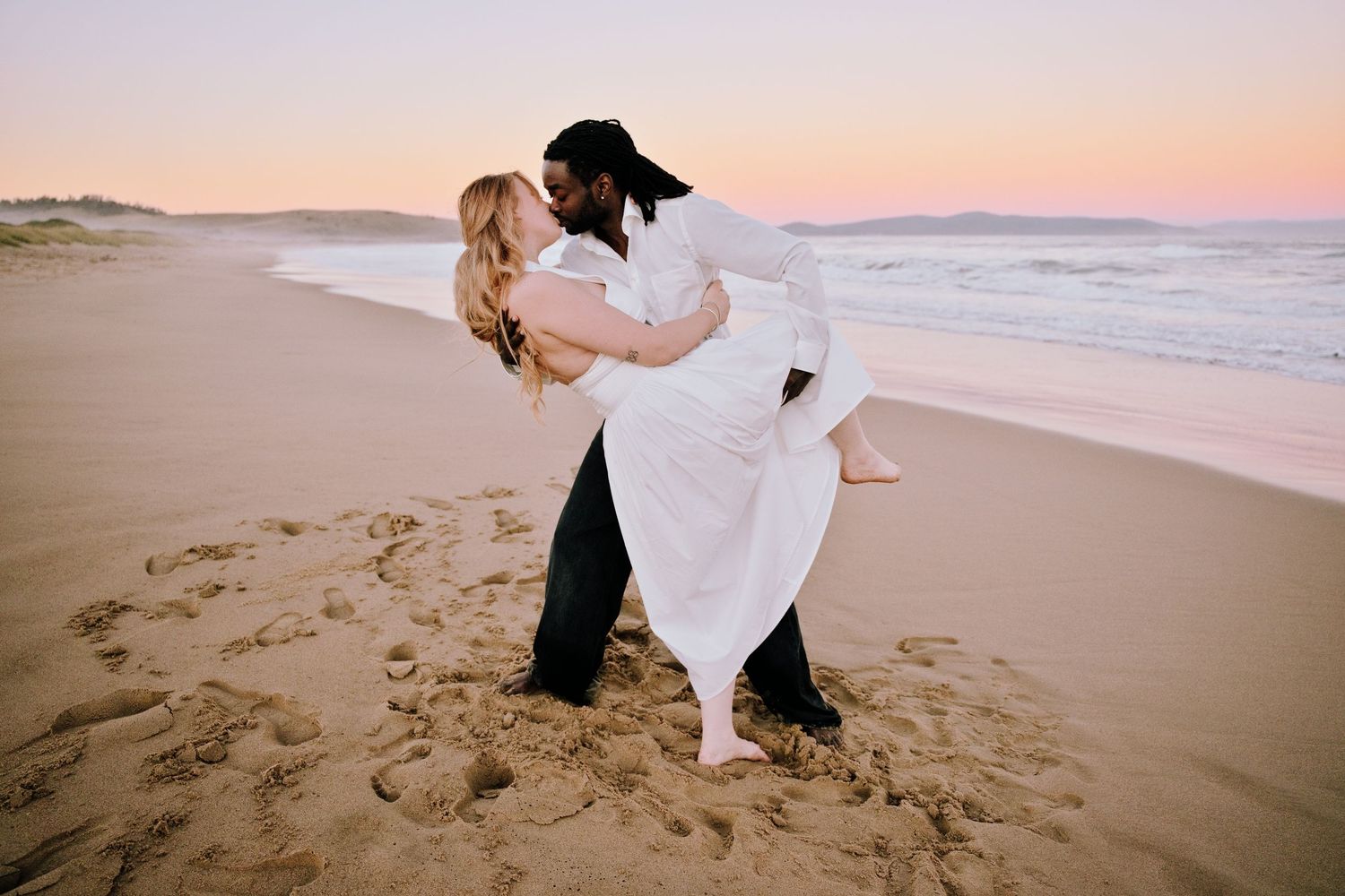 Couple sharing a romantic dip kiss on a sandy beach at sunset, wearing wedding attire.