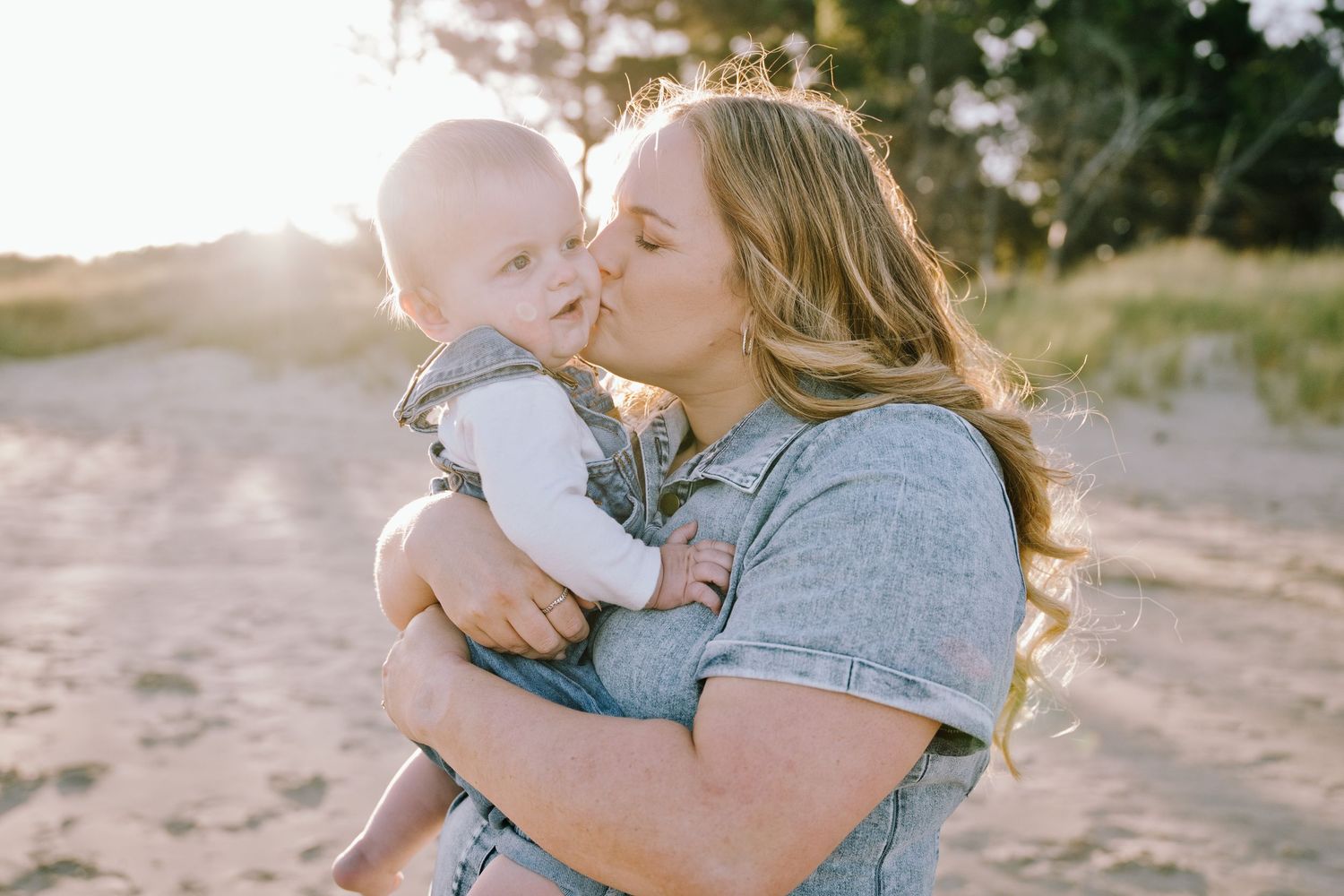 A mother gently kisses her baby outdoors in warm golden sunset light on a sandy path.