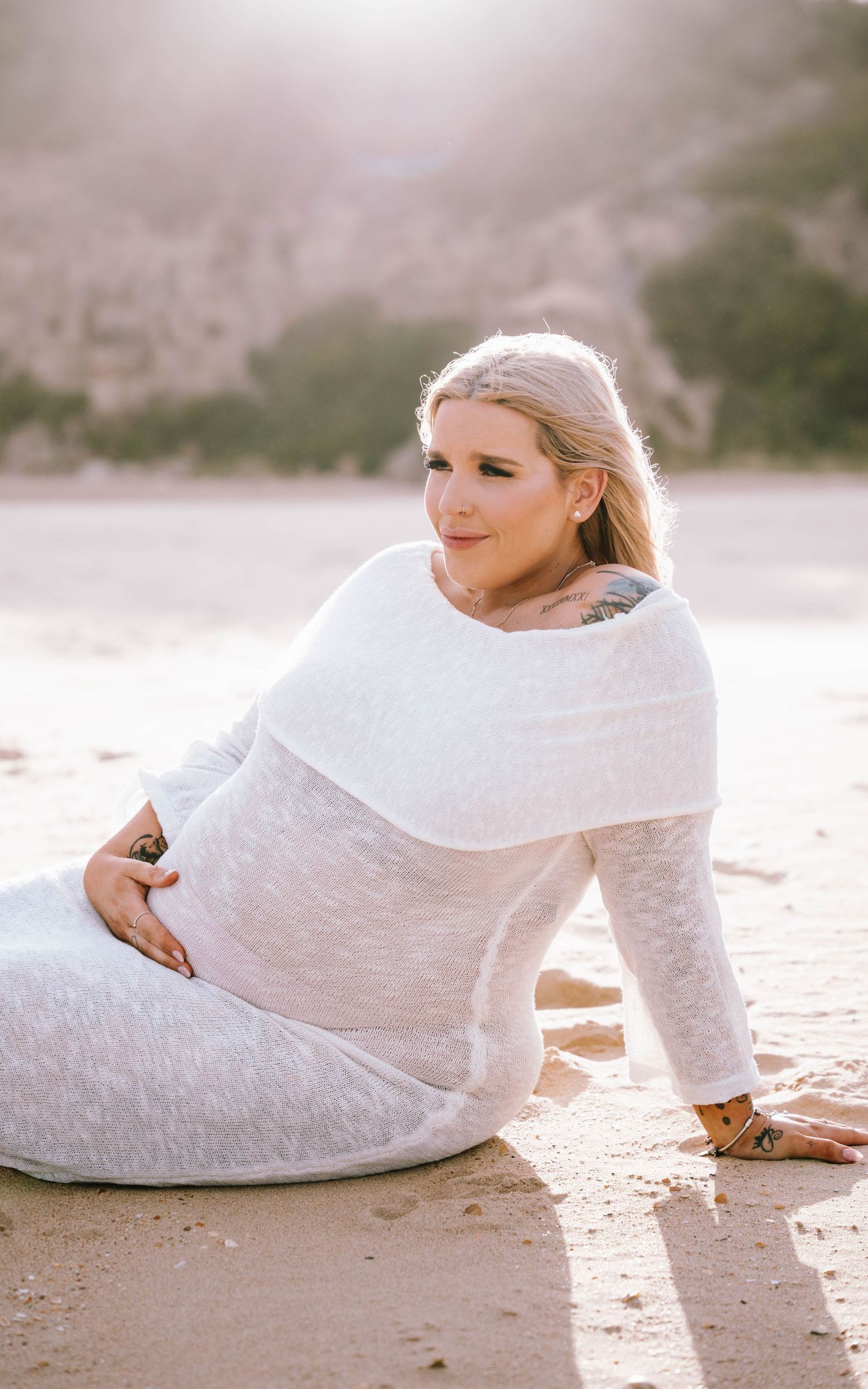 Serene maternity photo series showing someone in a white lace dress reclining on a sandy beach at sunset.