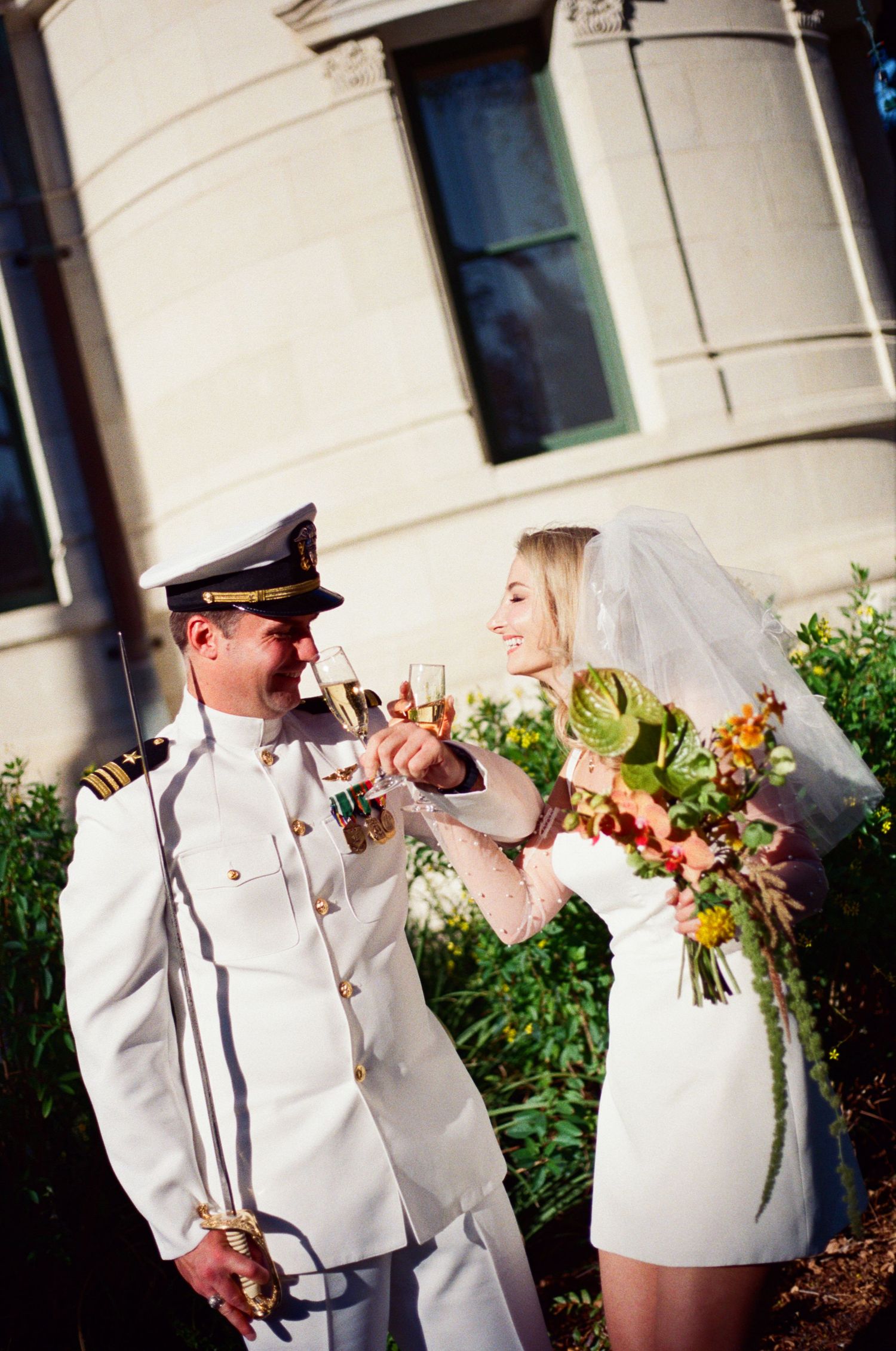 Military groom in white dress uniform and bride in mini dress toasting with champagne outside a historic building.
