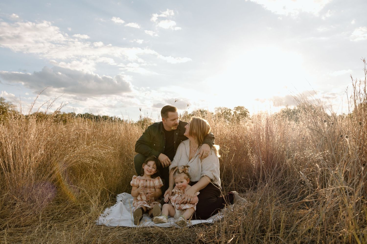 Family of four sitting together in tall golden grass field at sunset under dramatic cloudy sky.