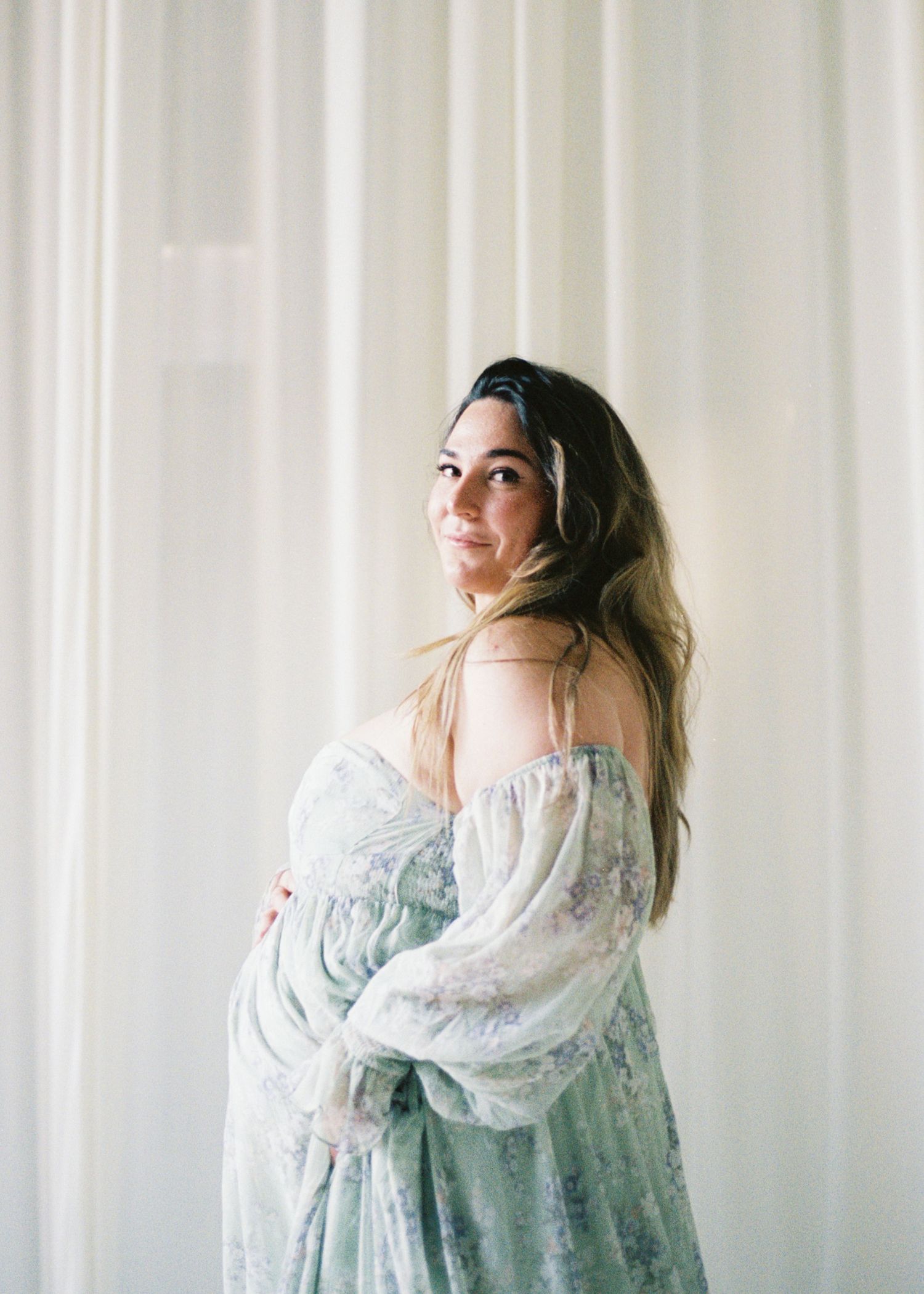 Pregnant woman in a floral mint dress poses by sheer white curtains during an elegant maternity photoshoot.