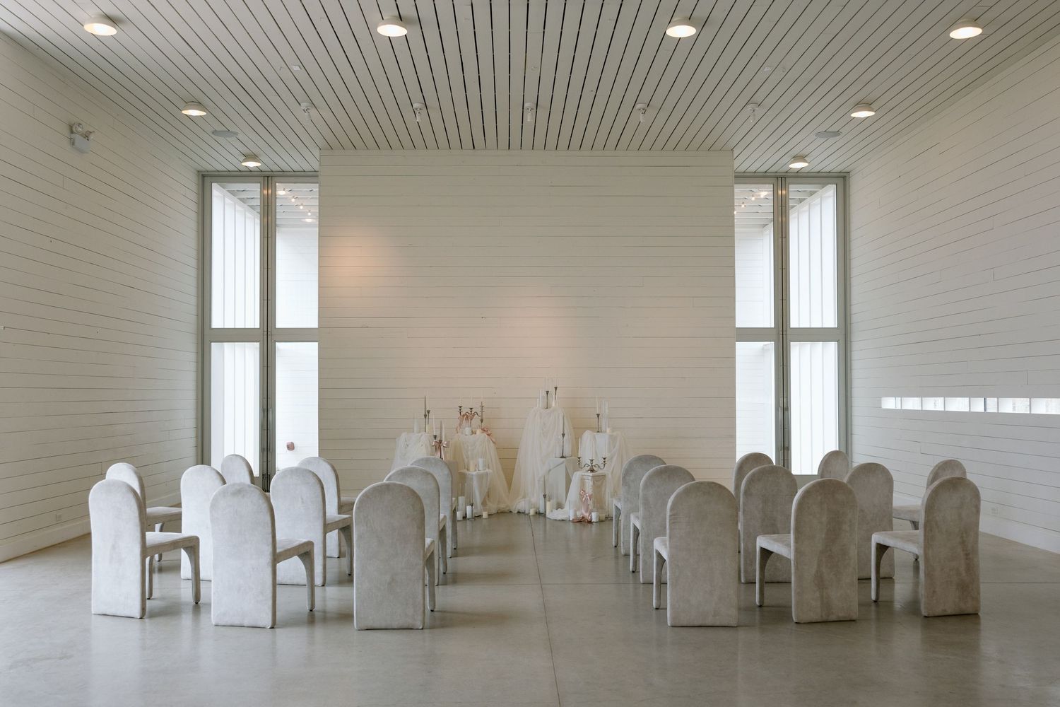 Minimalist white wedding ceremony venue with rows of elegant chairs and tall windows.