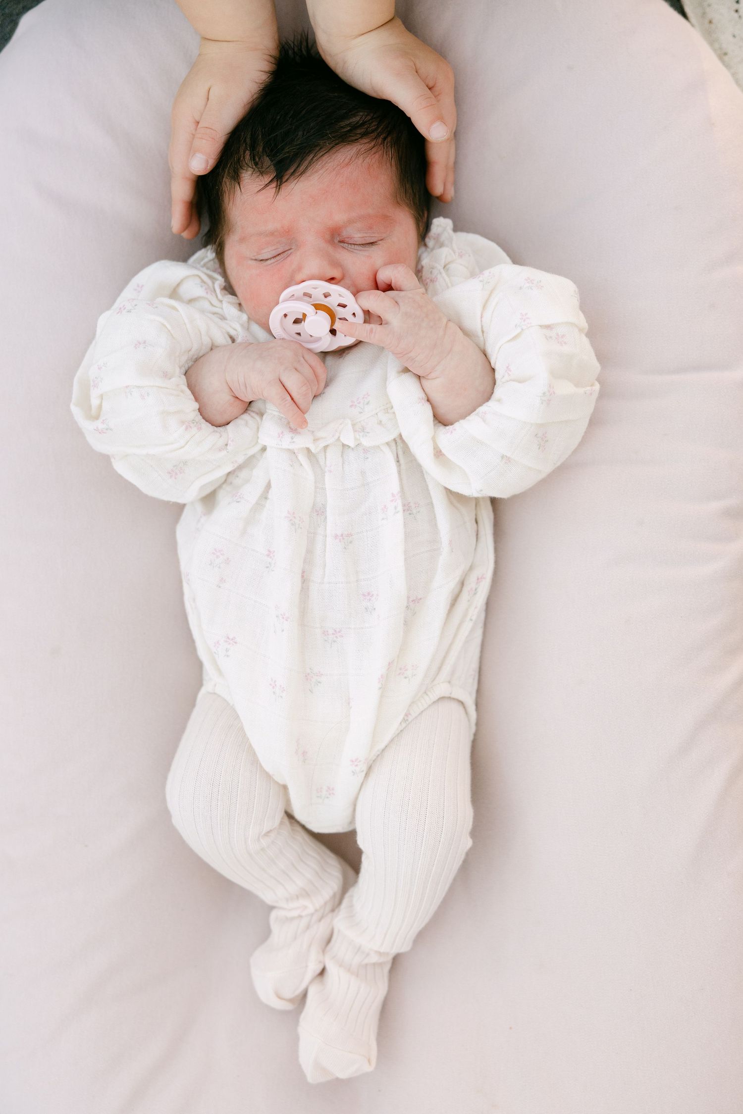 Newborn baby in white onesie sleeping peacefully with a pacifier while parent cradles head.