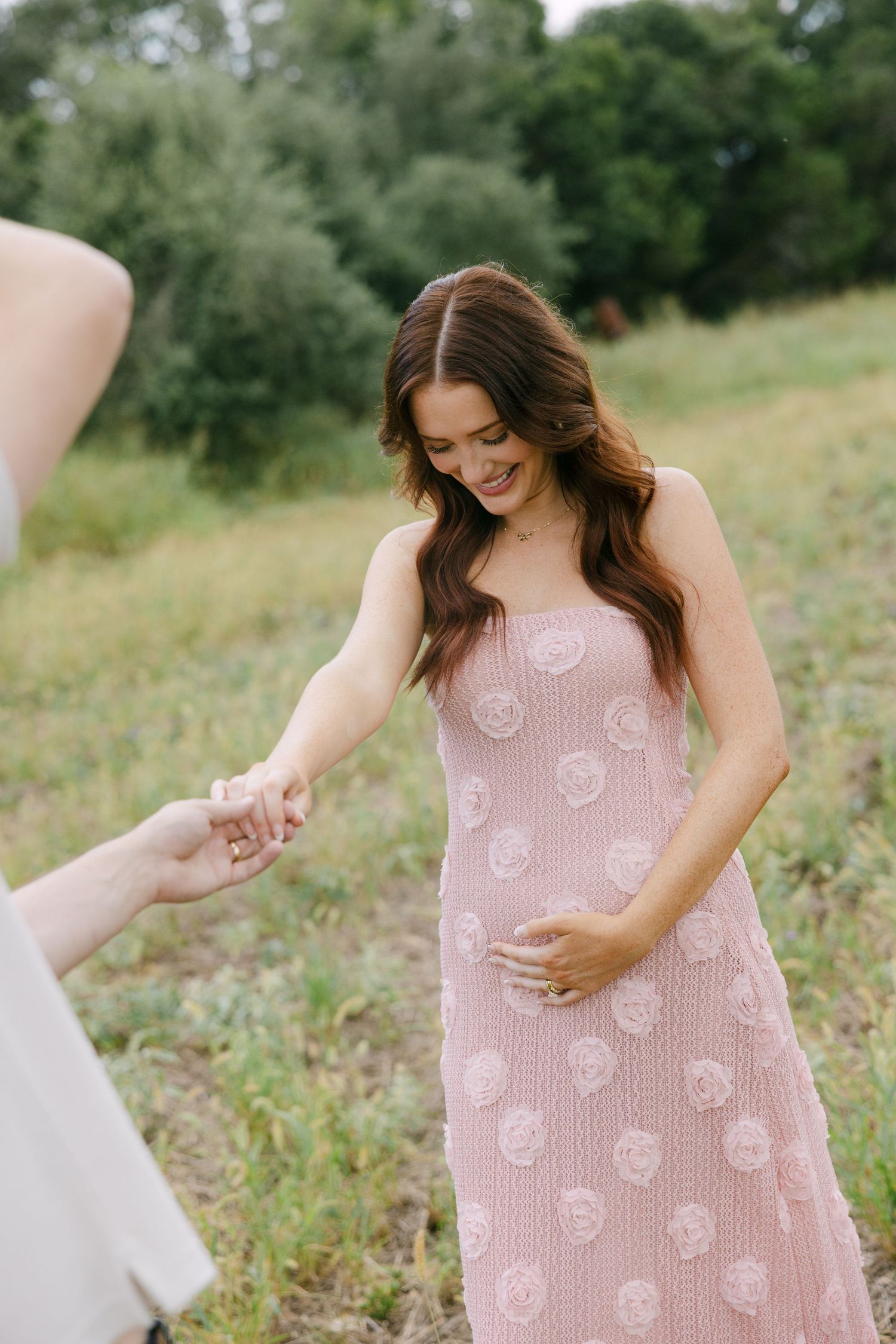 Smiling pregnant woman in a pink rosette dress holds hands with partner during an outdoor maternity session.