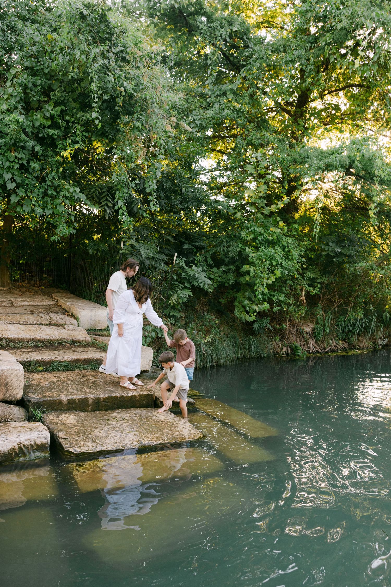 Mother in white dress helps young boy dip feet into clear green river water on stone steps surrounded by greenery.