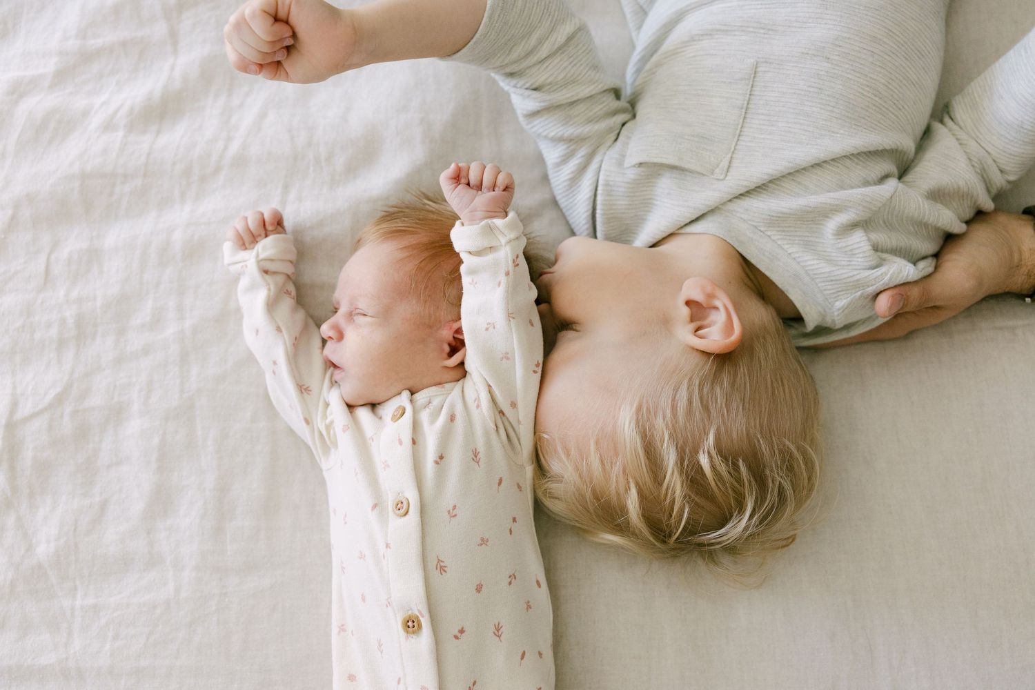 Newborn baby and toddler lying together on white bed, baby stretching arms.