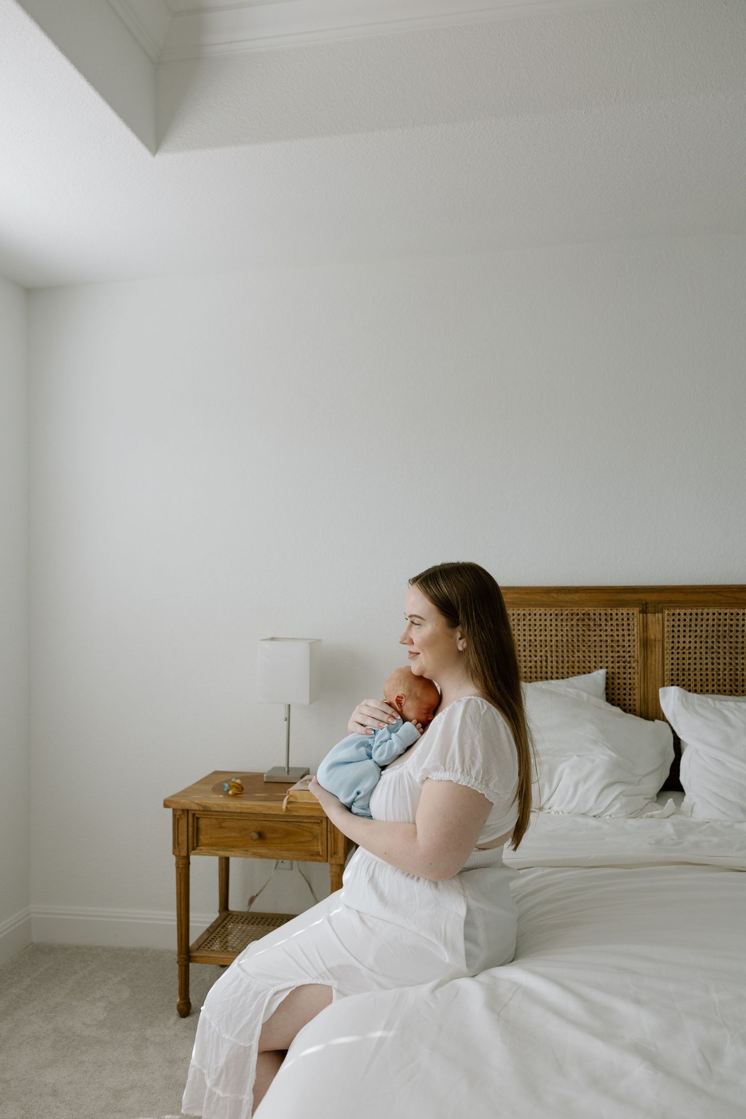 A woman in a white dress sits on a bed holding her newborn baby while gazing out a window.