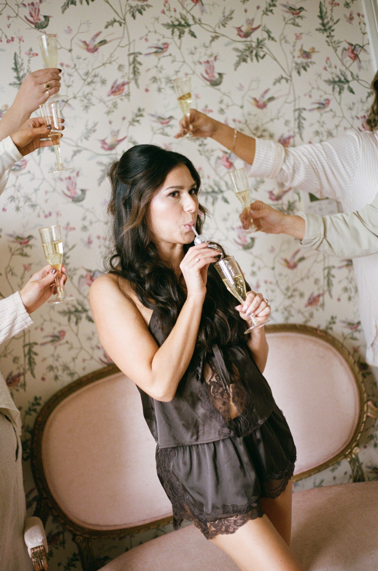 Bride in dark lingerie drinking champagne from a bottle surrounded by friends toasting at a floral wallpaper backdrop.