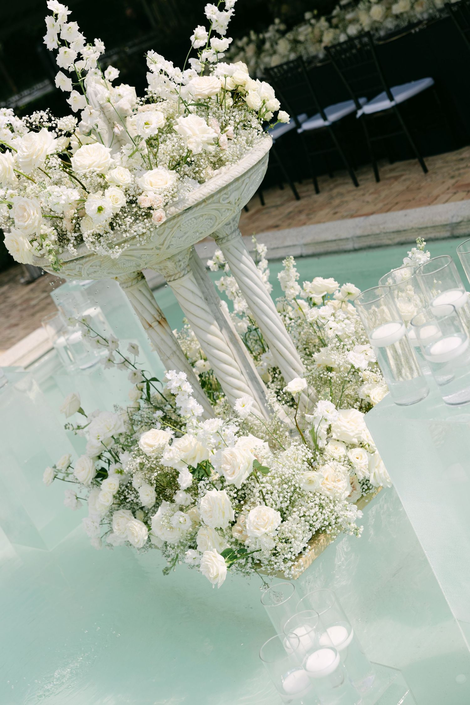 Stunning white floral arrangement with roses and baby's breath decorating a classical fountain at a wedding venue.