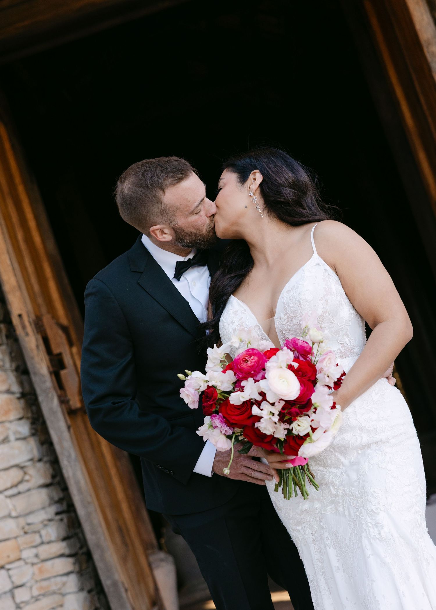 Bride and groom share a romantic kiss in a rustic doorway, bride holding a vibrant red and pink wedding bouquet.