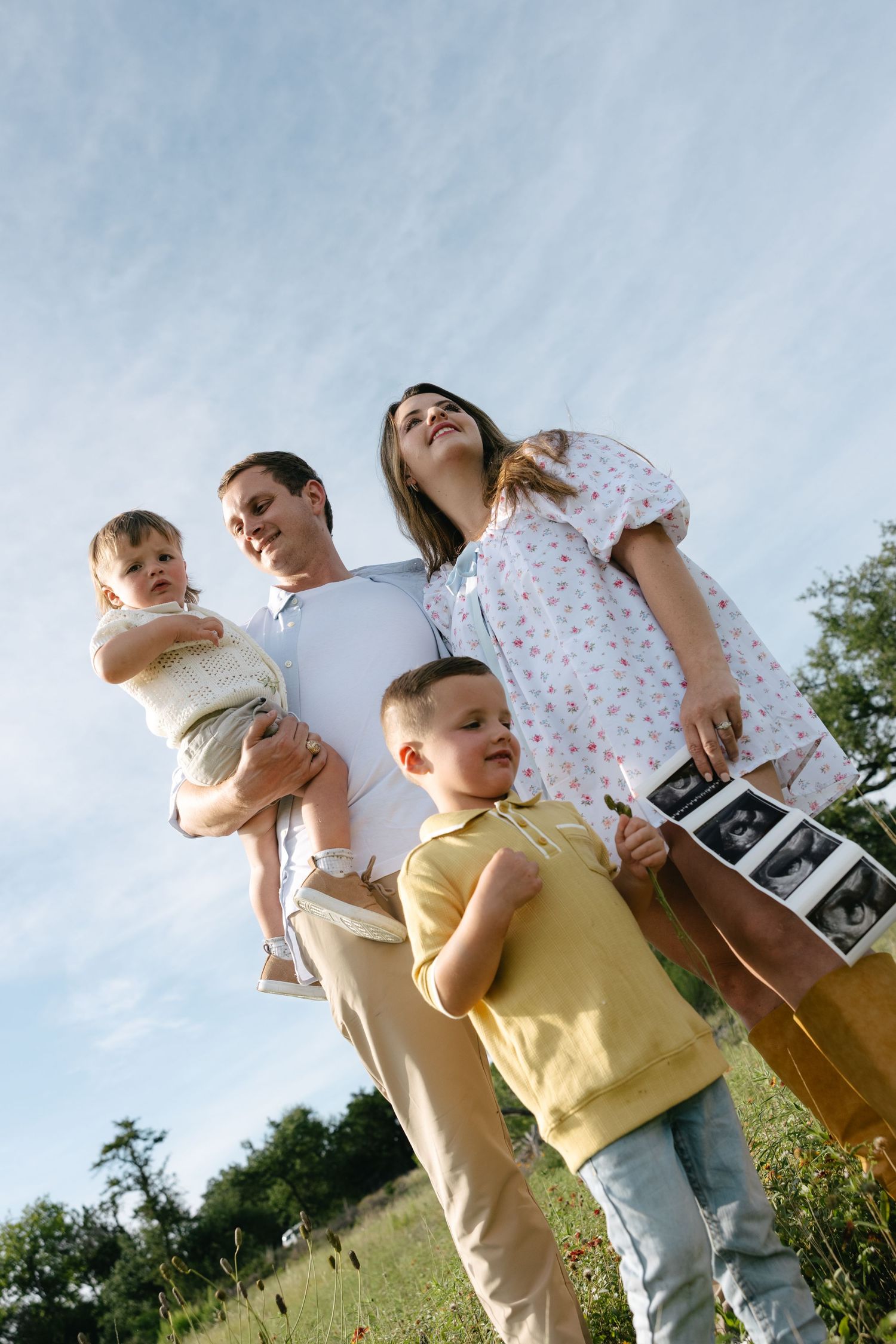 Happy family with pregnant mother holding ultrasound photos standing in sunny field.