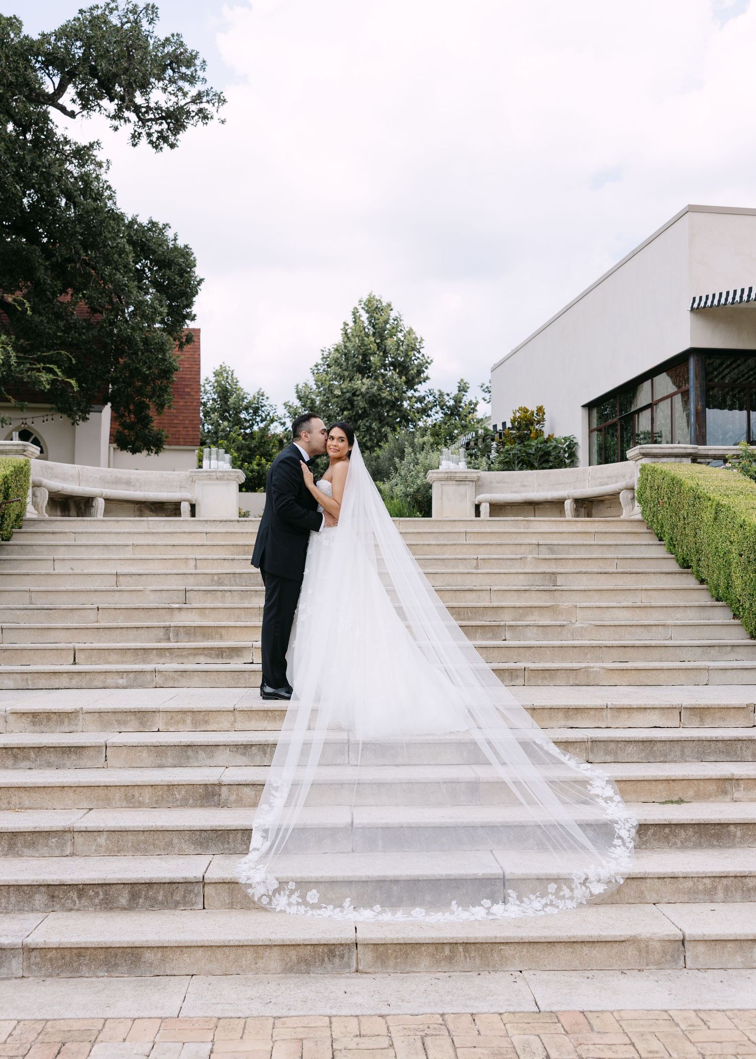 Bride and groom pose romantically on grand stone staircase, bride's cathedral lace veil trailing elegantly behind her.