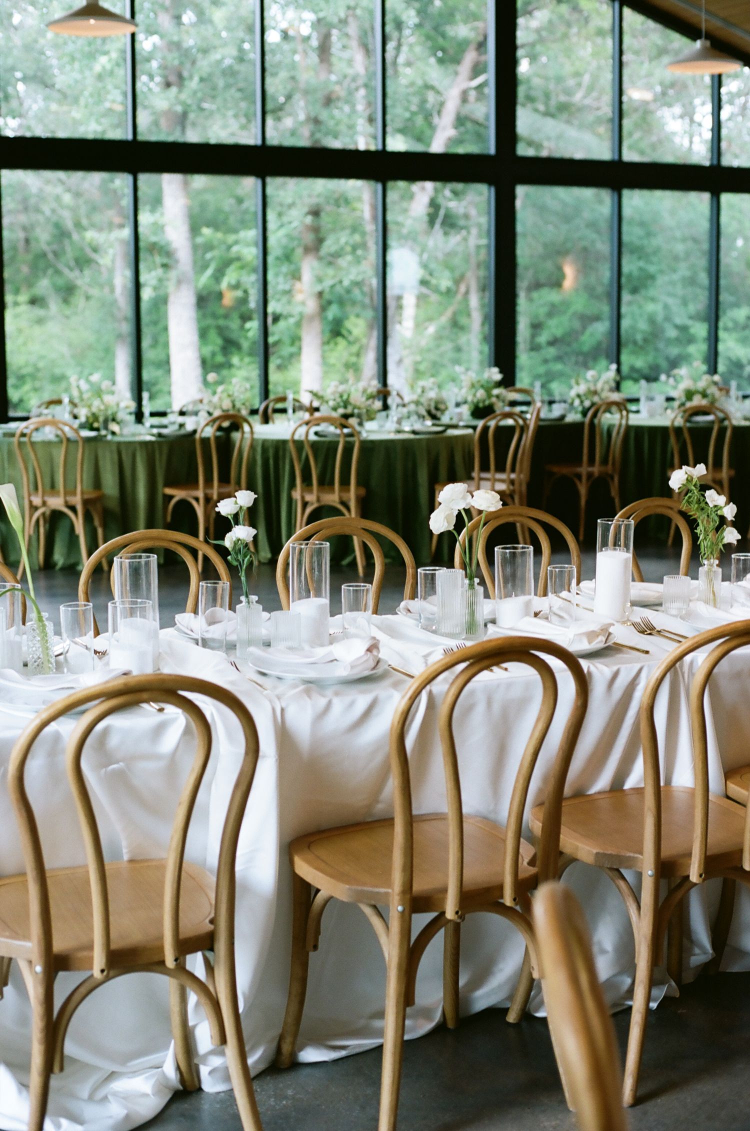 Elegant wedding reception with round tables, white linens, gold bentwood chairs, and white floral centerpieces by large windows.