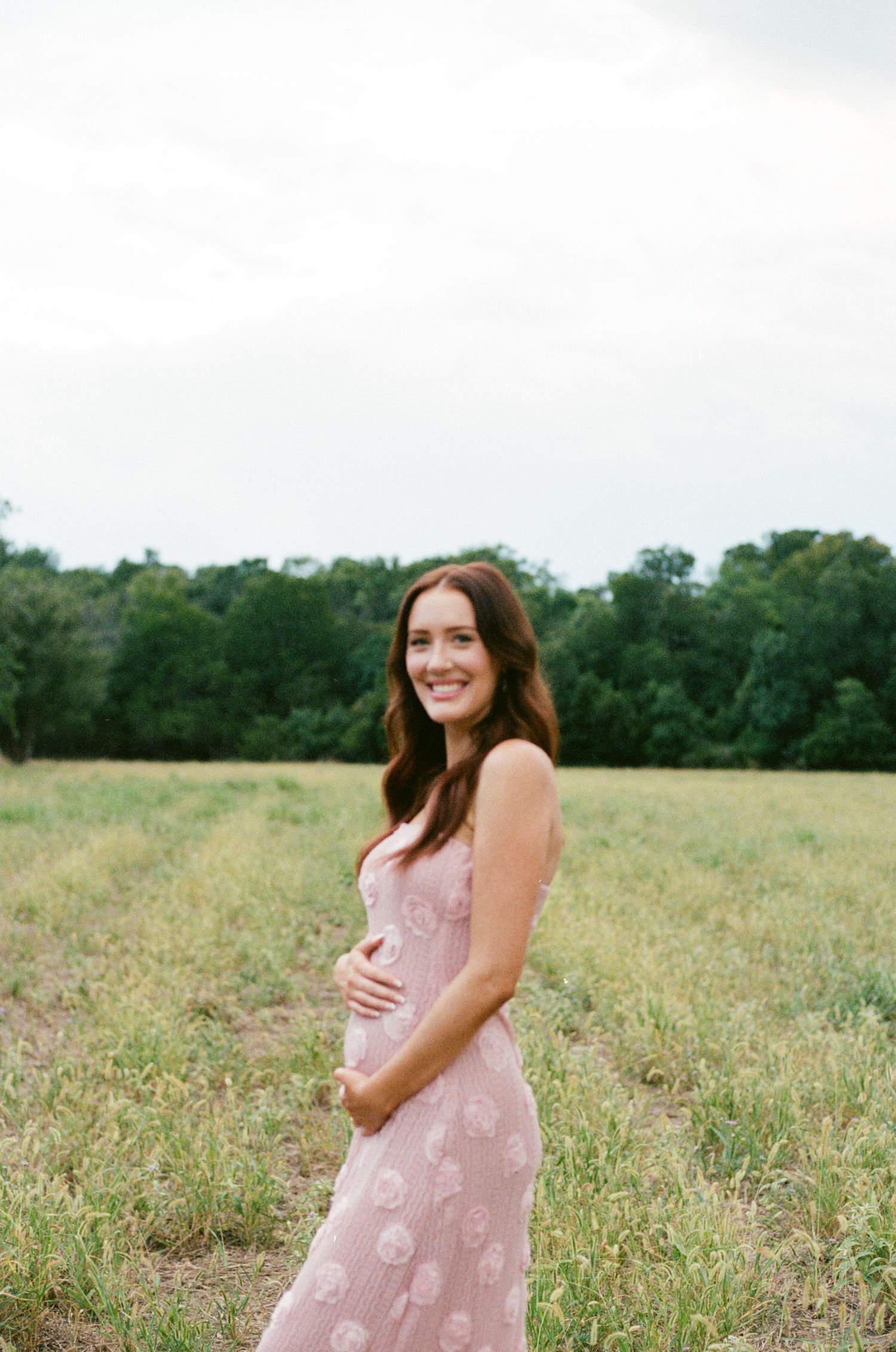 Smiling pregnant woman in pink floral dress cradling baby bump in green field.