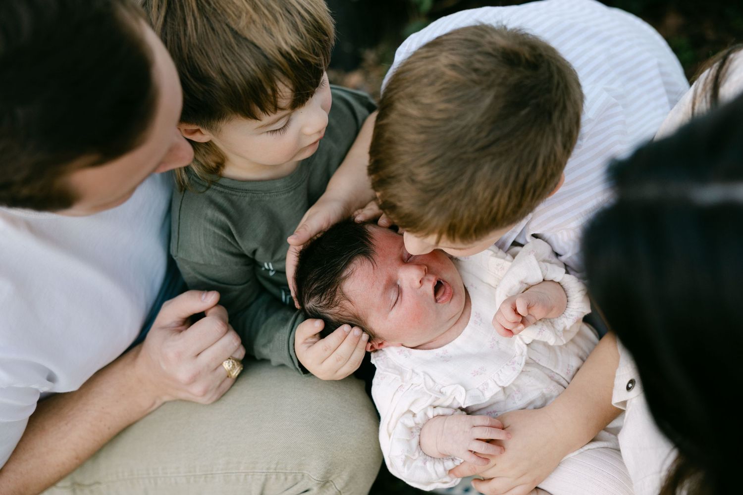 Overhead view of two young boys lovingly kissing a crying newborn baby held by parents.