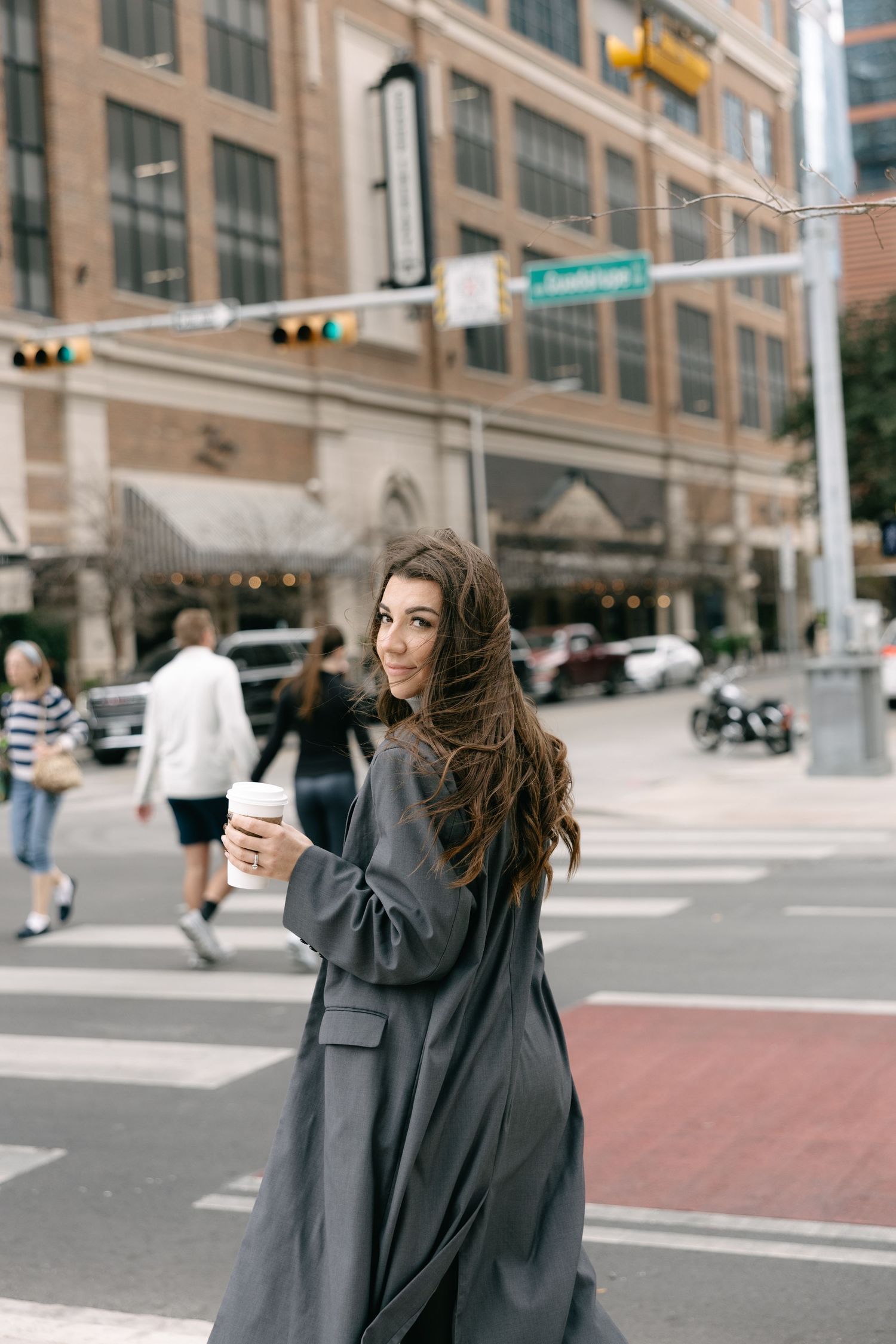 Stylish brunette woman in grey coat holding coffee cup while crossing a busy urban street intersection.