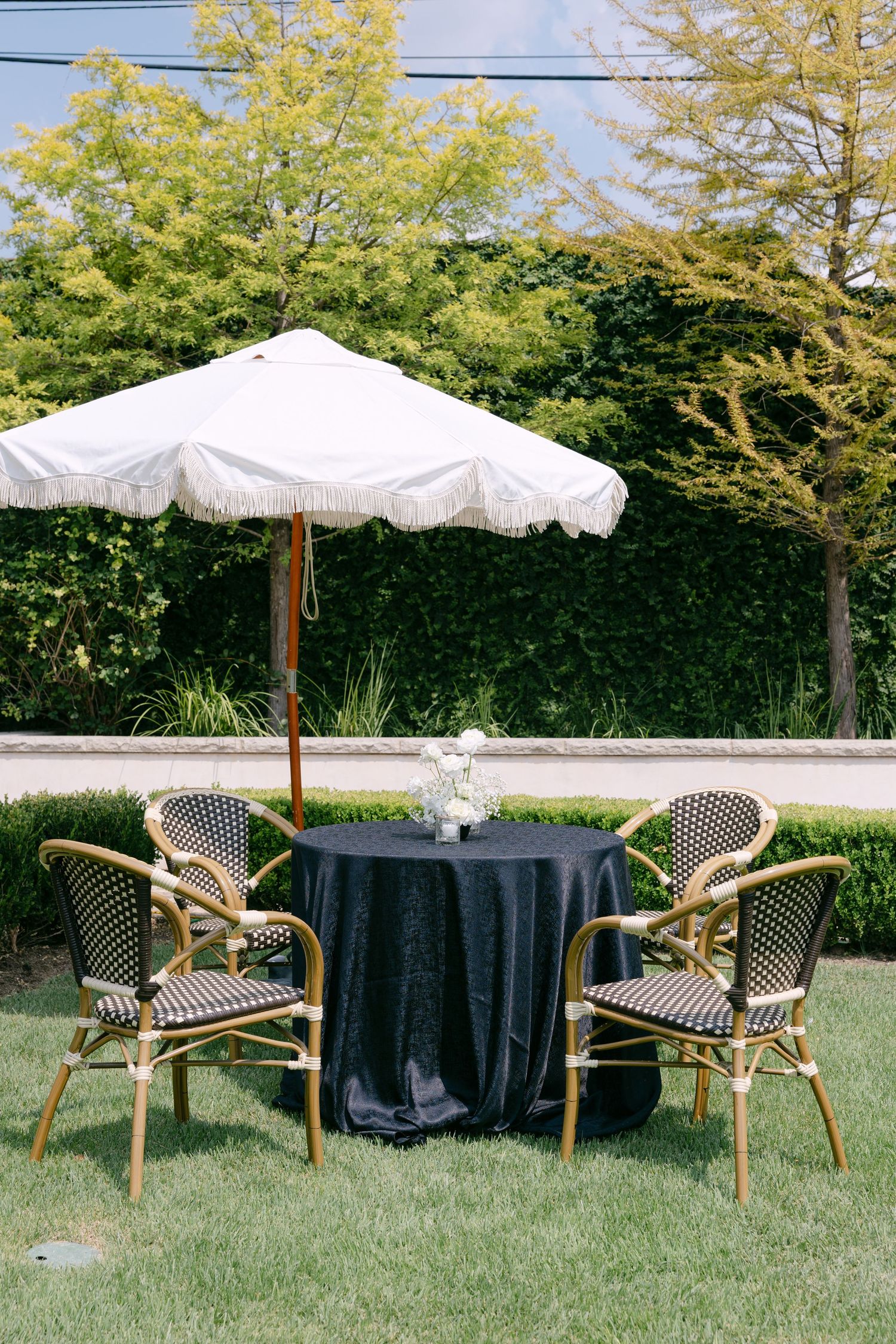 Outdoor wedding reception seating with a navy tablecloth, wicker chairs, and a white fringed patio umbrella on green lawn.