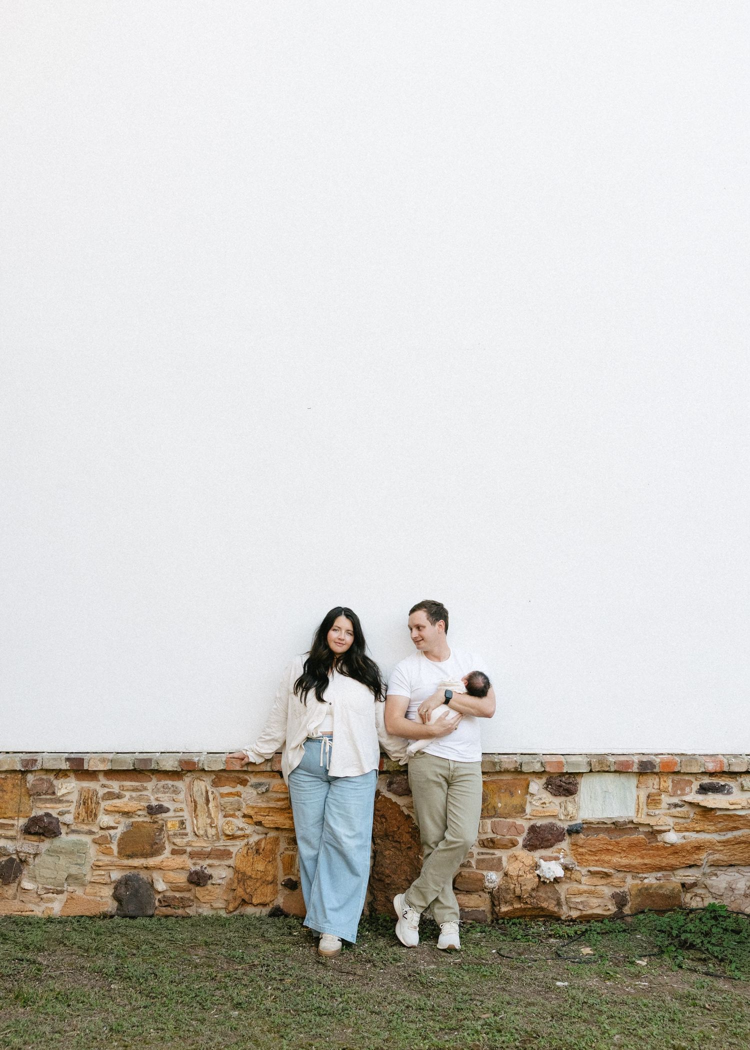 A young couple stands against a stone wall outdoors, smiling, with the man holding a newborn baby.