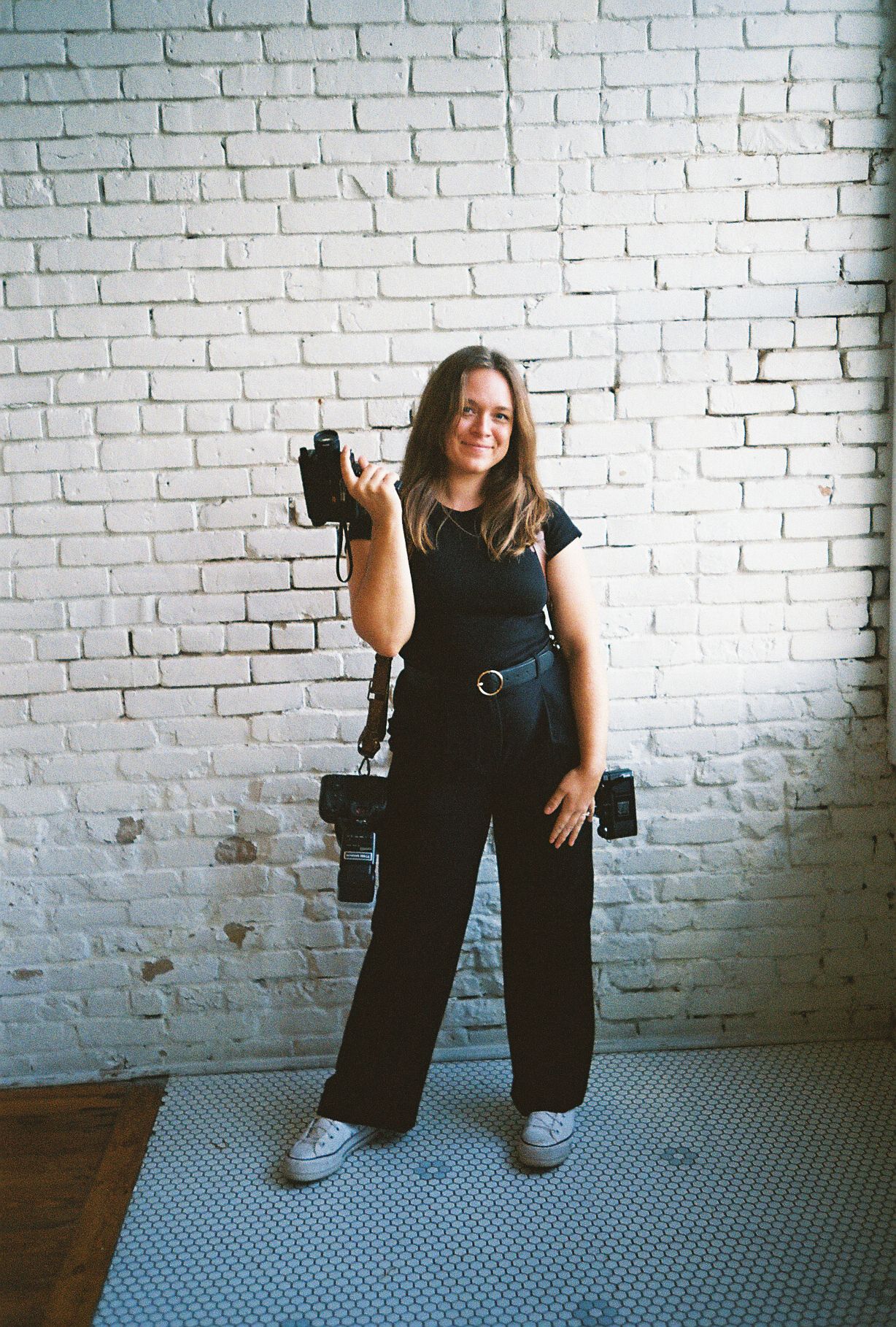 Smiling woman in black outfit holding cameras in front of white brick wall.