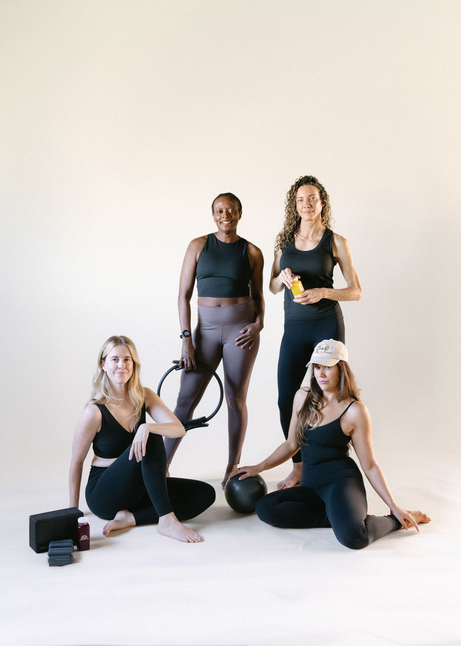 Four women in black athletic wear pose with fitness props including a pilates ring, yoga block, and medicine ball in a studio.