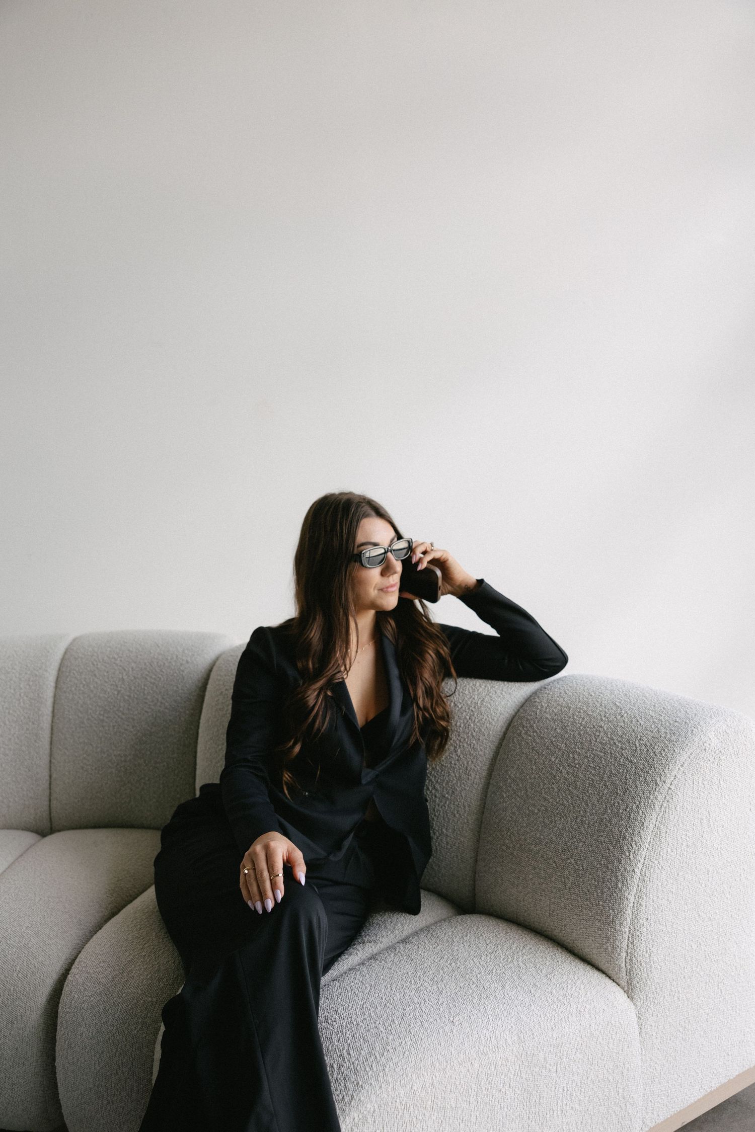 Stylish woman in black suit wearing sunglasses sitting on gray boucle sofa.