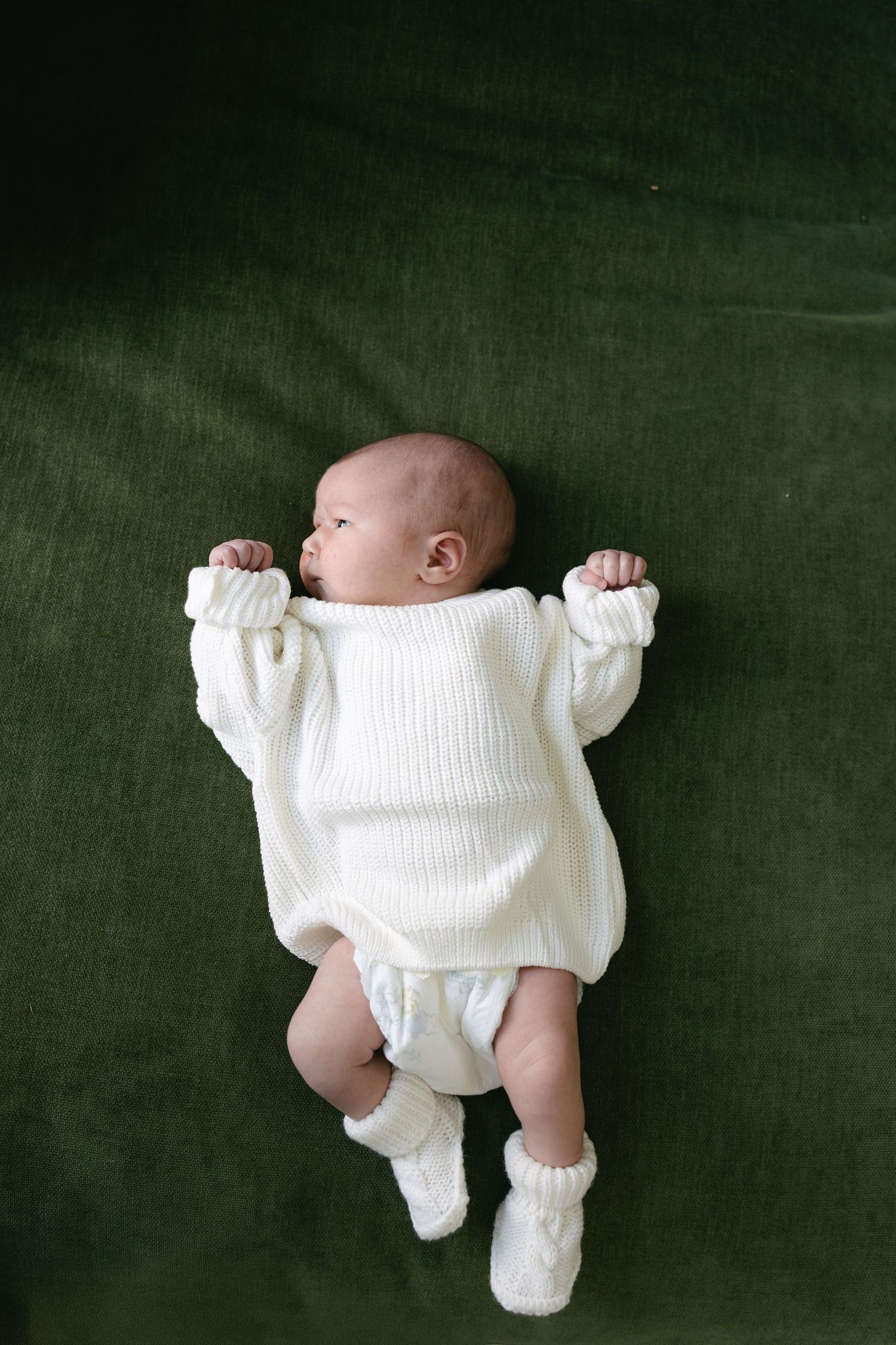 Newborn baby in a white knit romper and booties lying on a green velvet surface.
