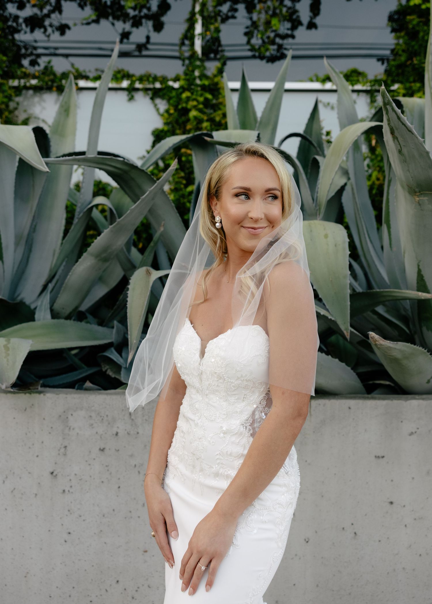 Smiling blonde bride in a fitted lace wedding gown and veil posing before large agave plants.