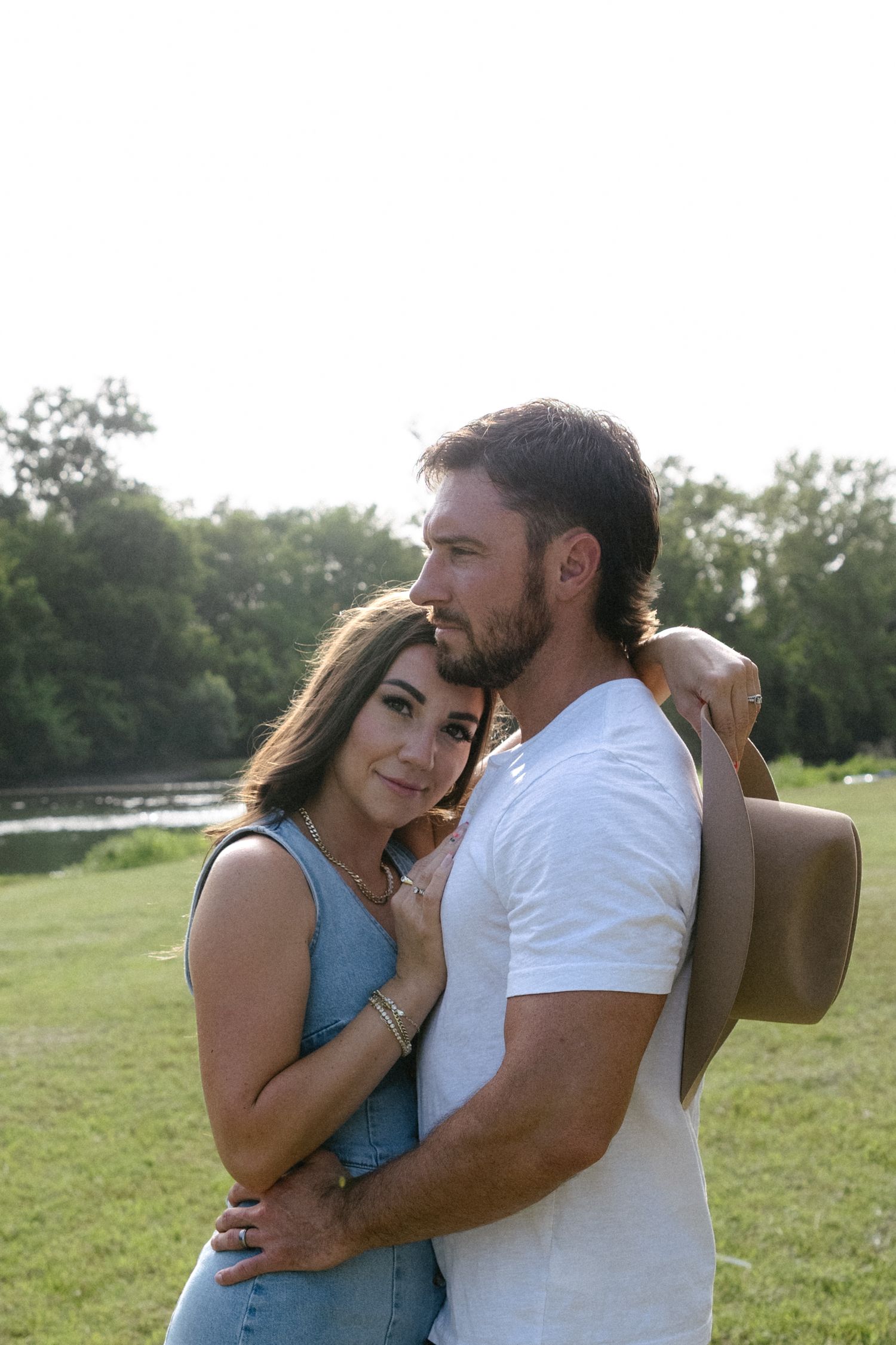 Romantic couple embraces outdoors near a river, woman in denim dress resting head on bearded man holding a hat.