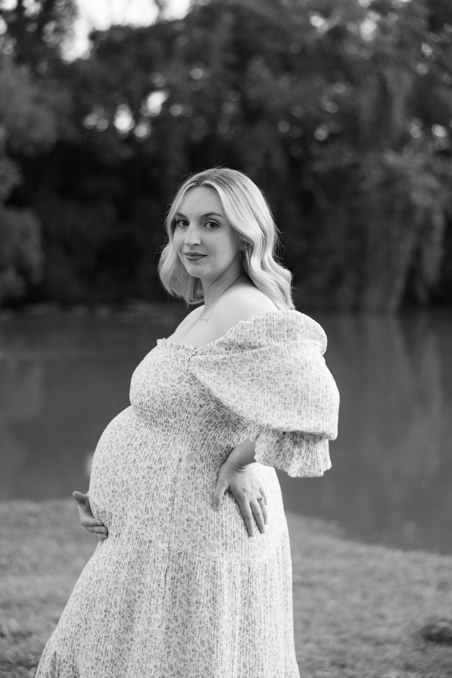 Black and white portrait of pregnant blonde woman in floral dress near water.