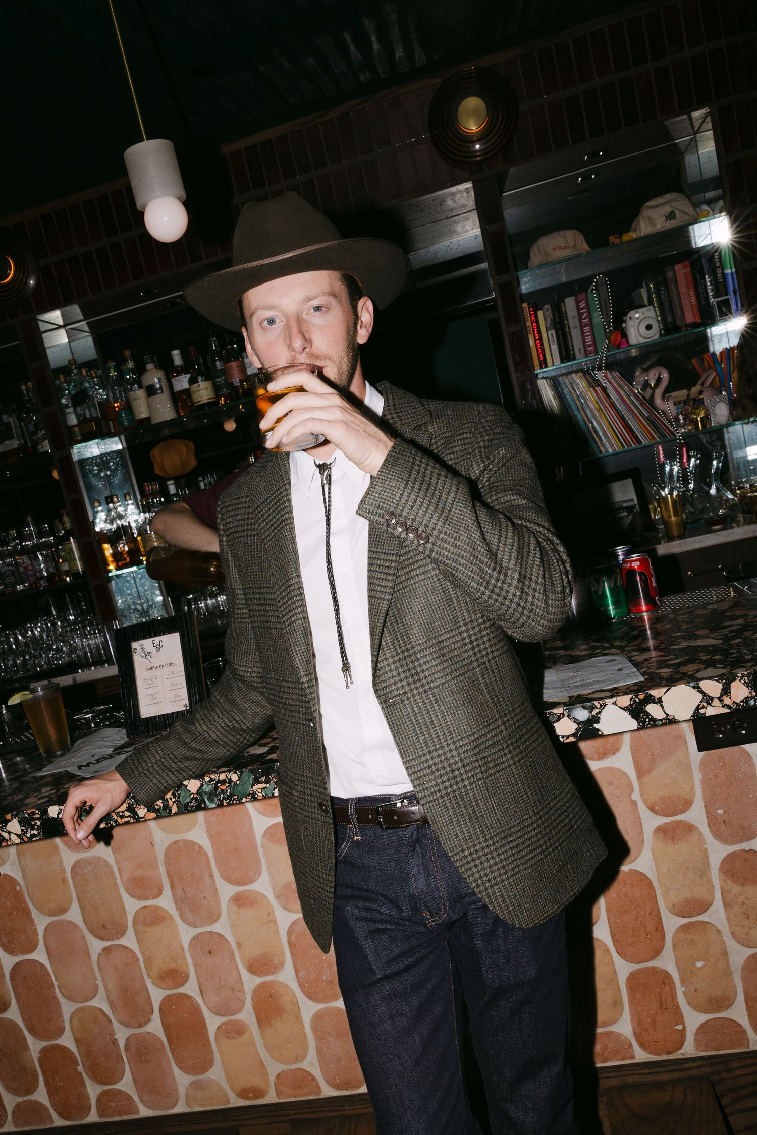 Stylish wedding guest in a cowboy hat and tweed blazer enjoys a drink at a dimly lit bar.
