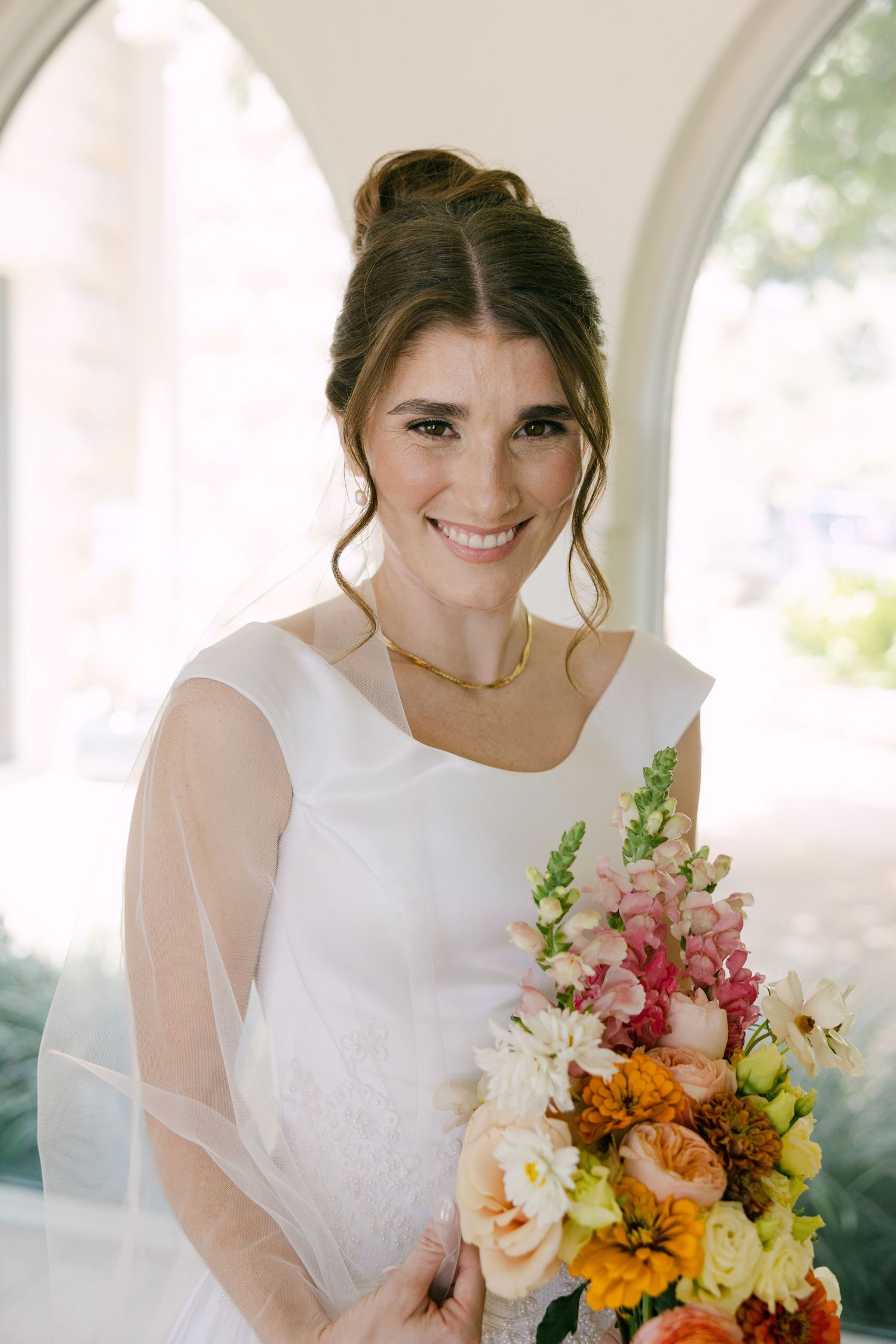 Smiling bride in a modern white wedding dress holds a colorful bouquet of orange, pink, and yellow flowers.
