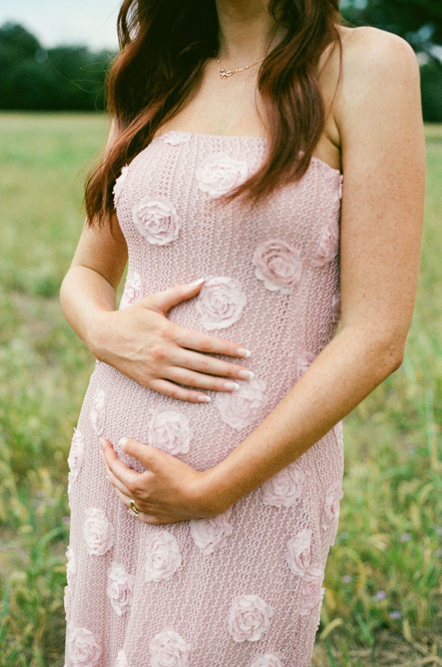 Close-up of a pregnant woman in a pink rosette dress cradling her baby bump during an outdoor maternity shoot.