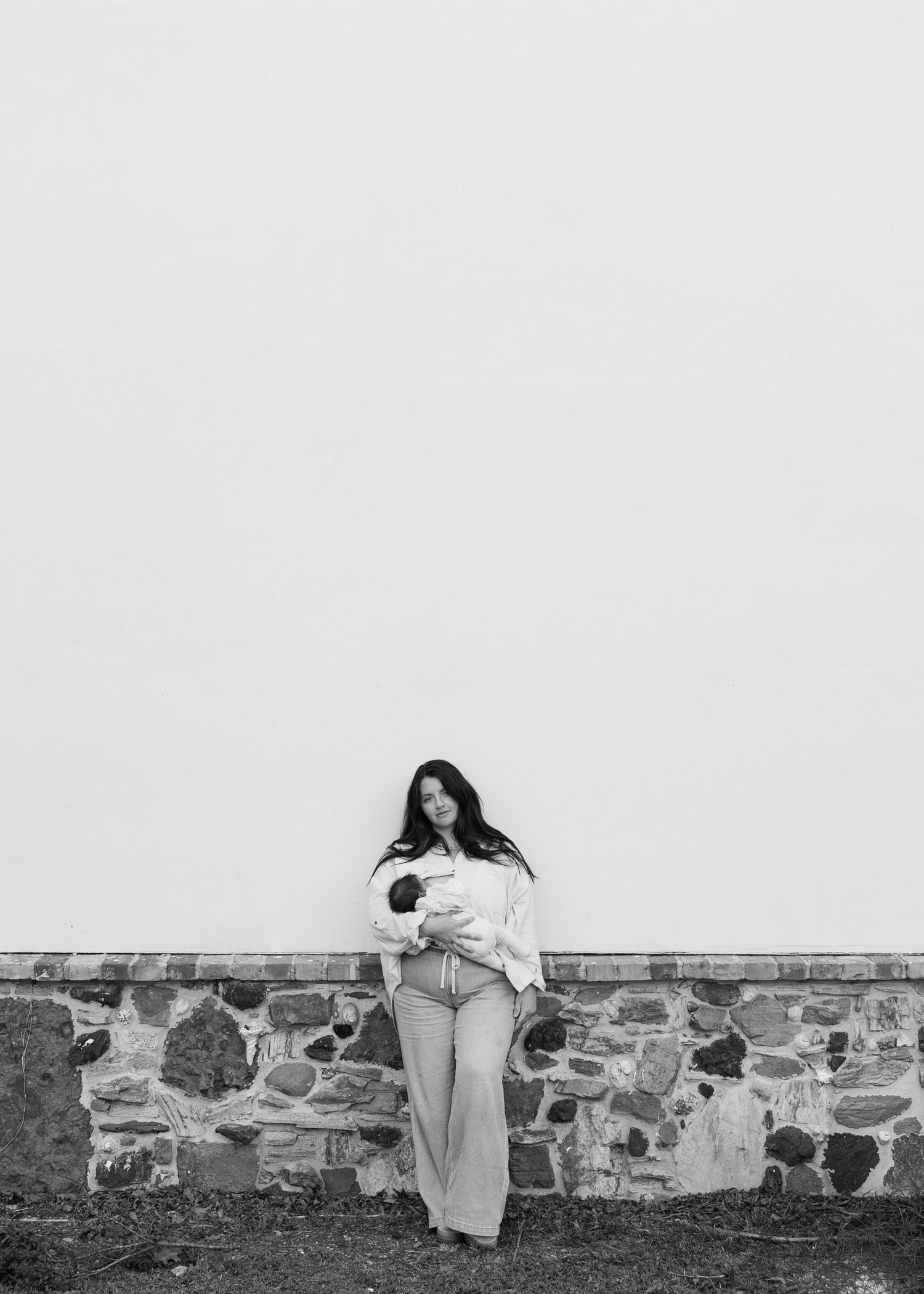Black and white portrait of a woman with long dark hair holding a newborn baby against a stone wall.