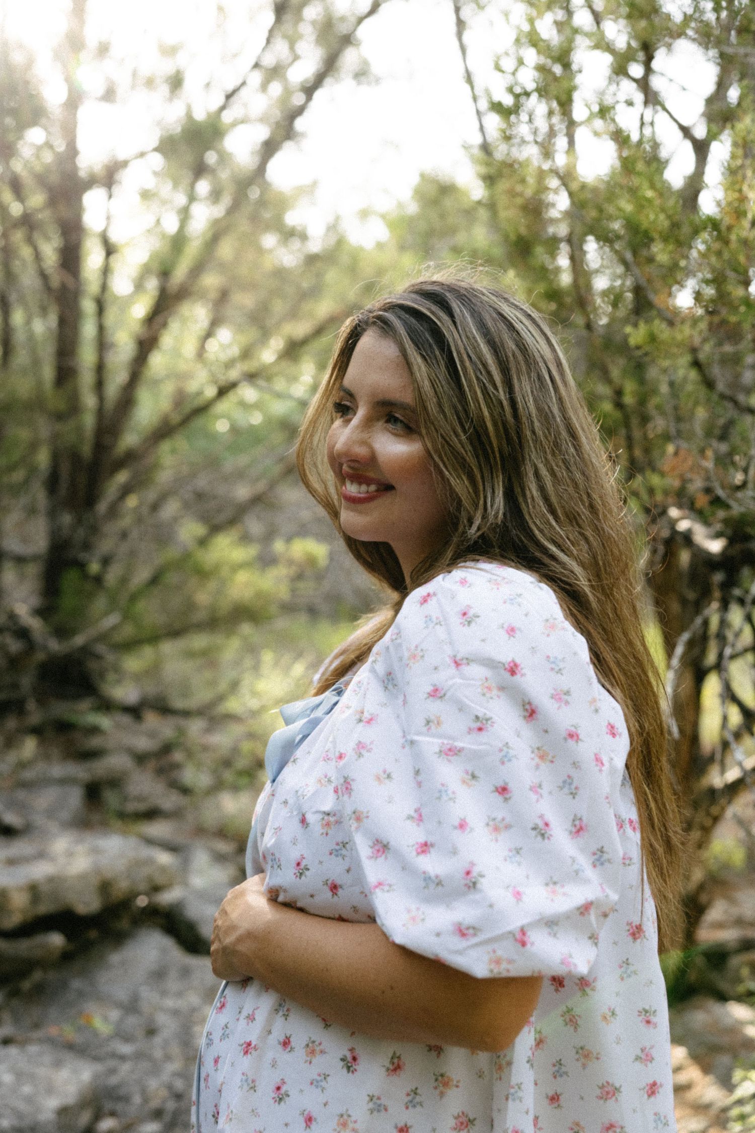 Smiling pregnant woman in floral dress holding baby bump in a sunlit forest setting.