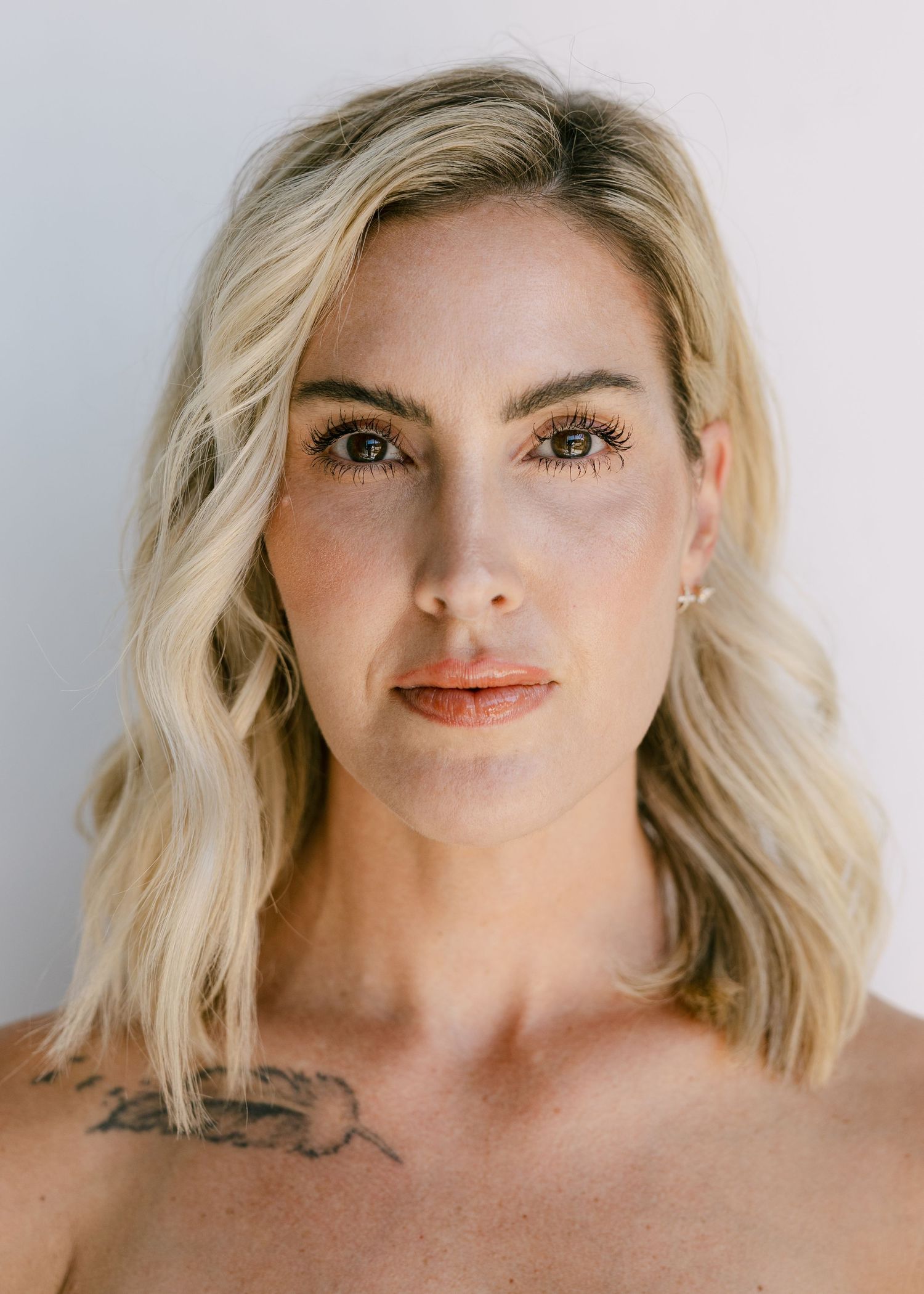 Blonde woman with tattoo on chest poses for a natural beauty portrait against white background.