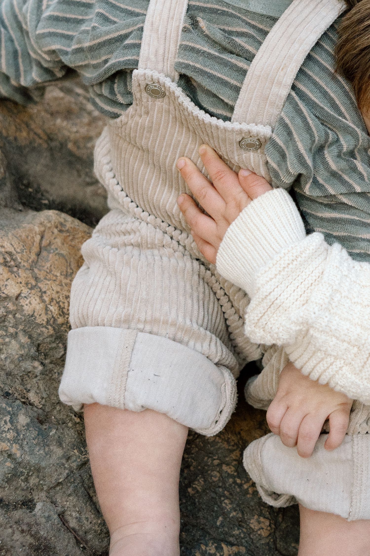 Close-up of baby wearing cream knit sweater and beige corduroy overalls sitting on rocks outdoors.