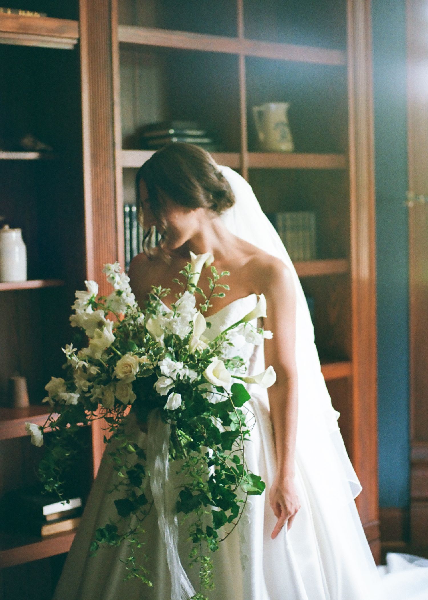 Bride in strapless white gown smelling a lush white and green cascading bouquet indoors near a wooden bookshelf.