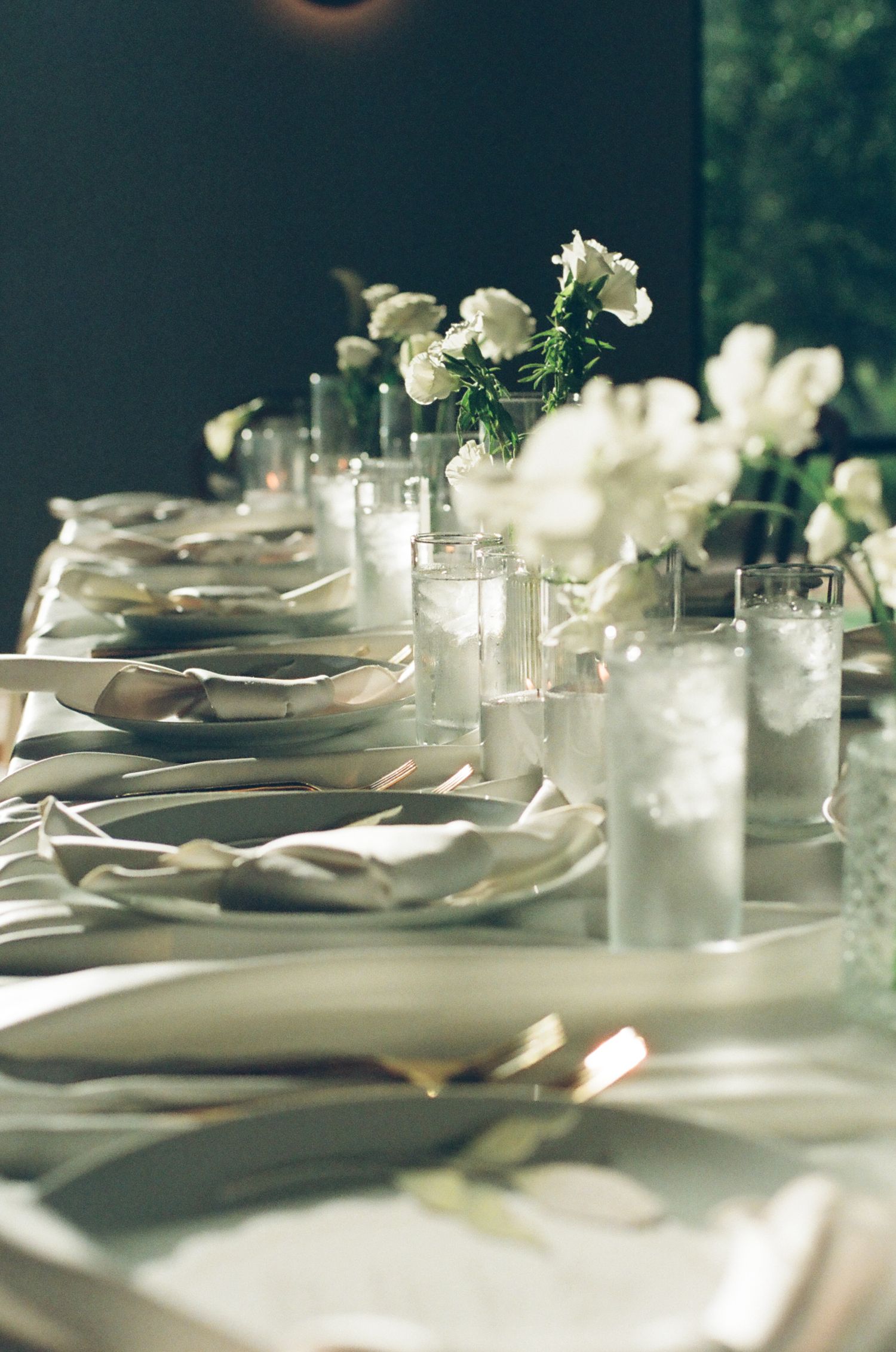 Elegant wedding tablescape with white floral arrangements, candles, gold chargers, and draped napkins in soft moody lighting.